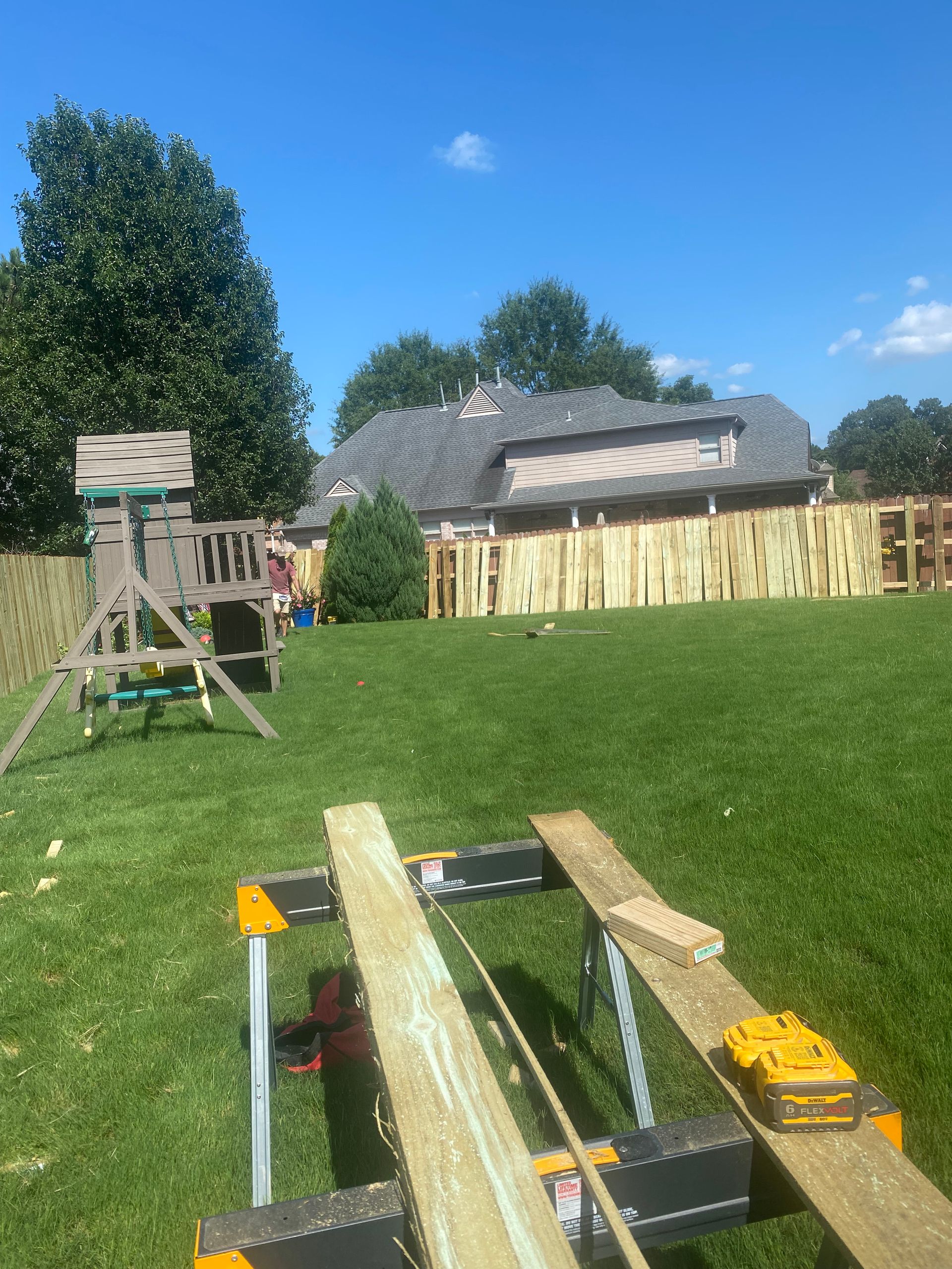 A backyard scene with a wooden fence being built. Boards rest on a sawhorse in the foreground, with a swing set and house in the background.