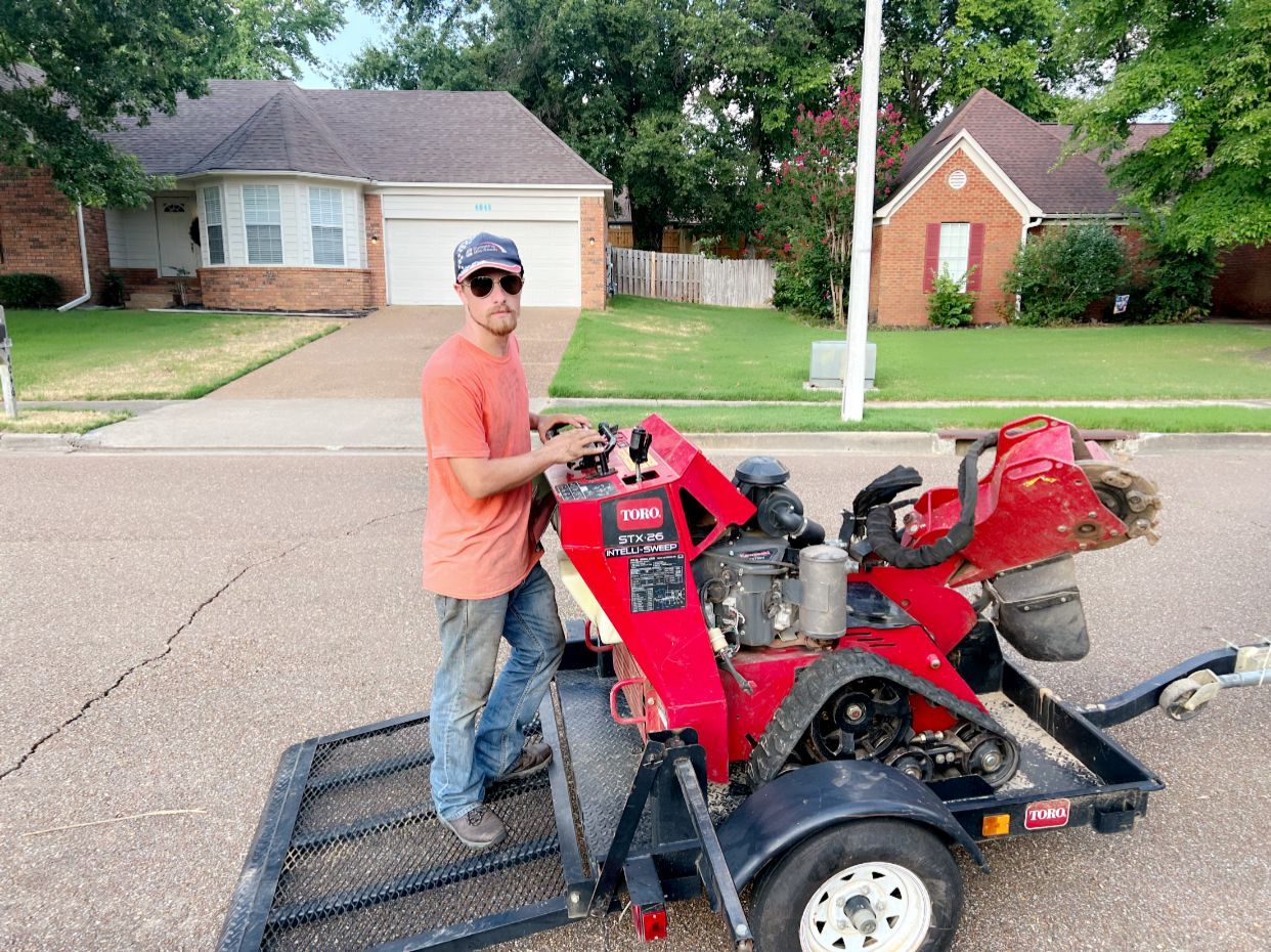 Man operating a red stump grinder on a trailer in front of a suburban house.