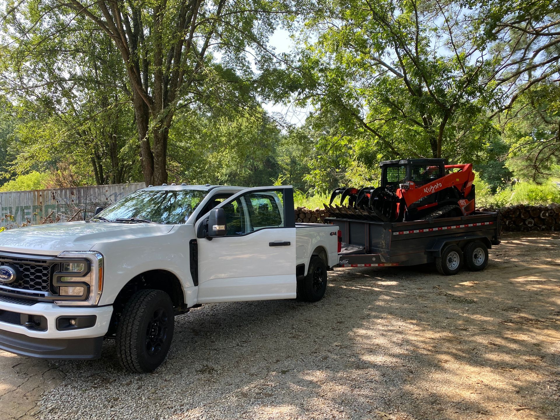 White Ford truck with door open, towing a trailer carrying an orange skid steer in a wooded area.