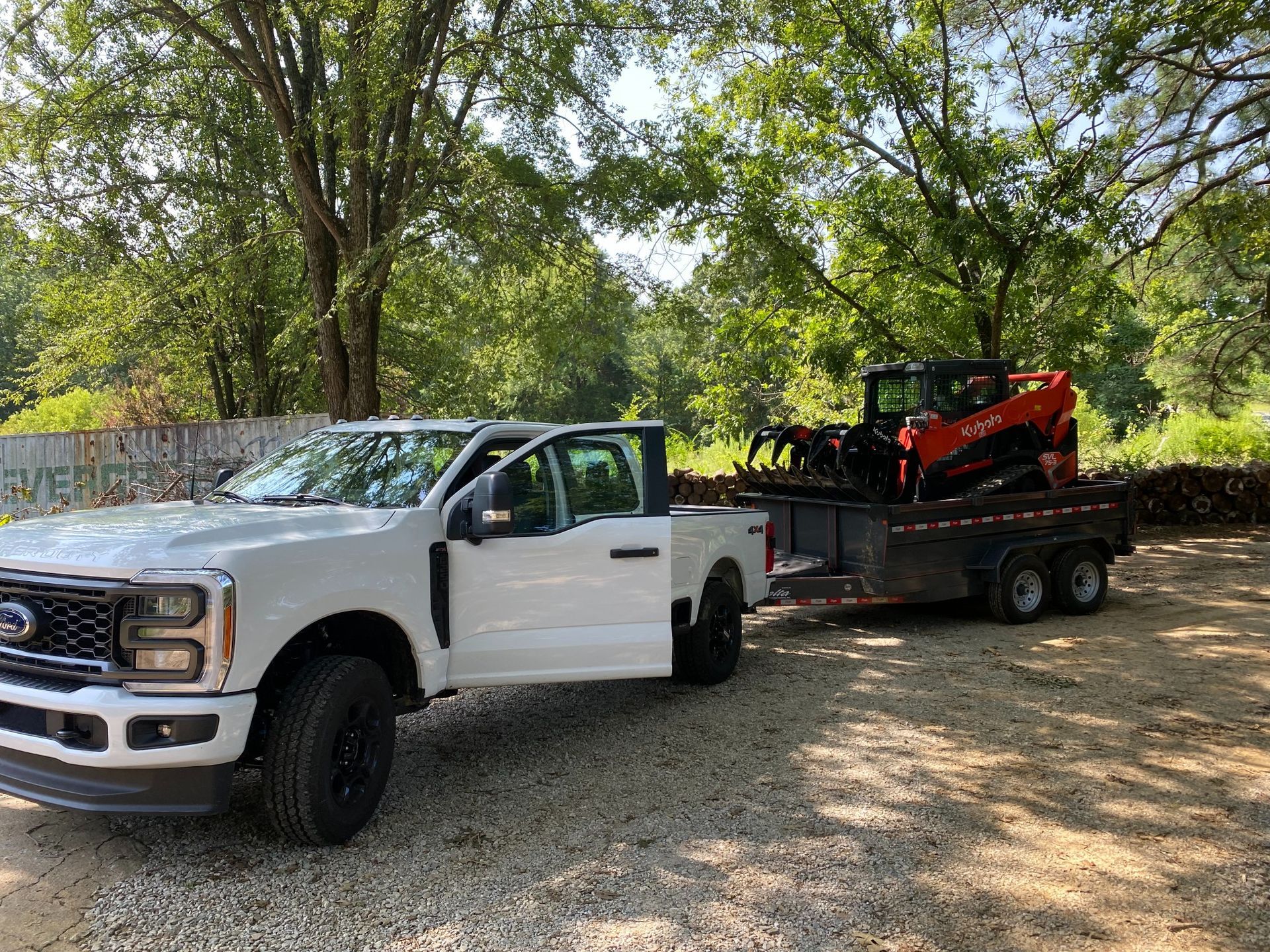 White Ford truck with open door pulling a trailer with an orange tractor on it, parked on gravel by trees.