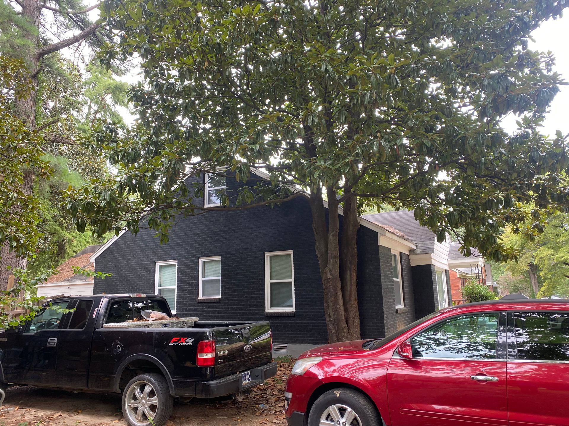 Black house with white-trimmed windows, a truck, and a car parked in front of a large tree.