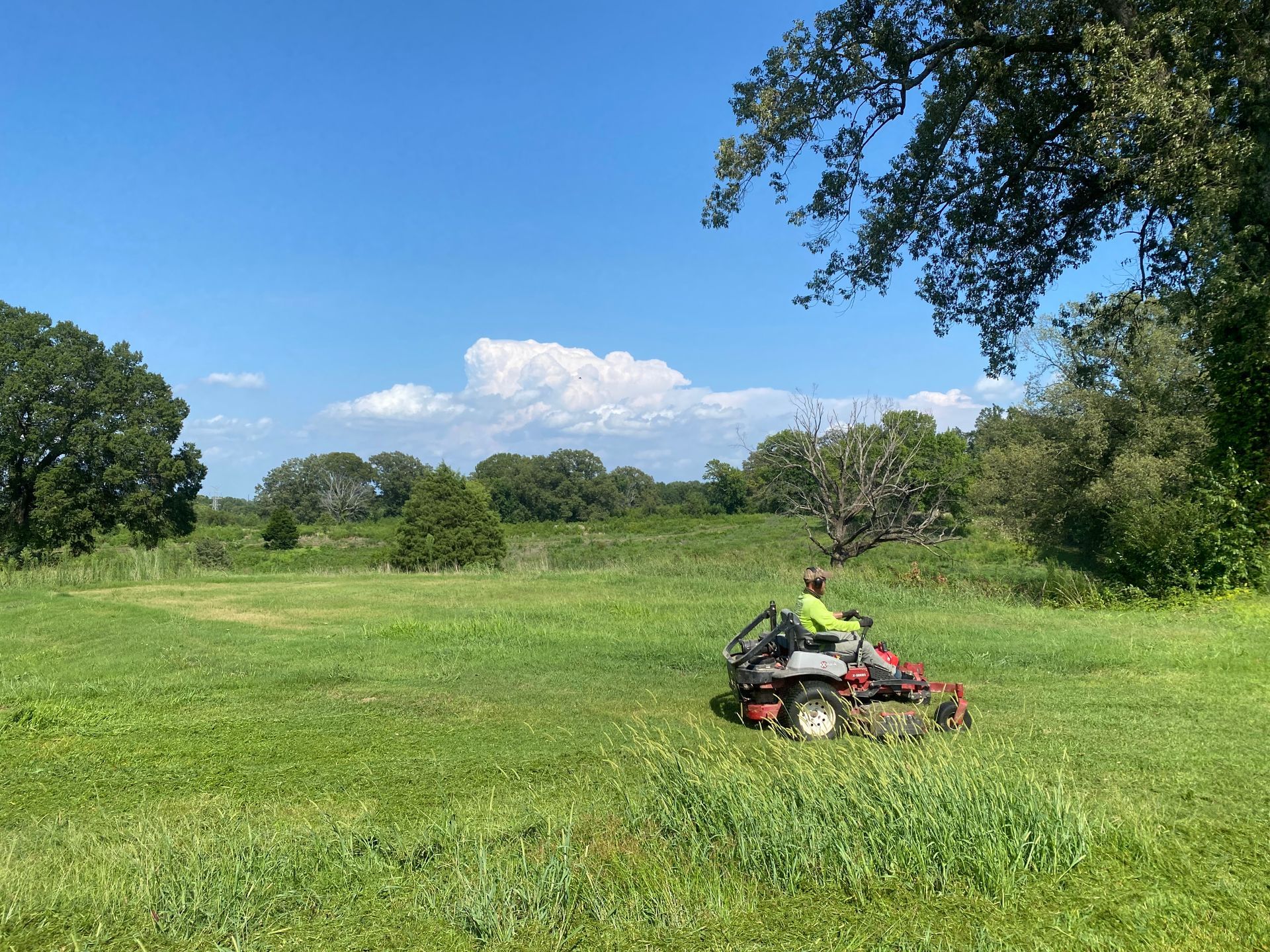 Person on a riding lawnmower cuts grass in a green field under a blue sky with trees.