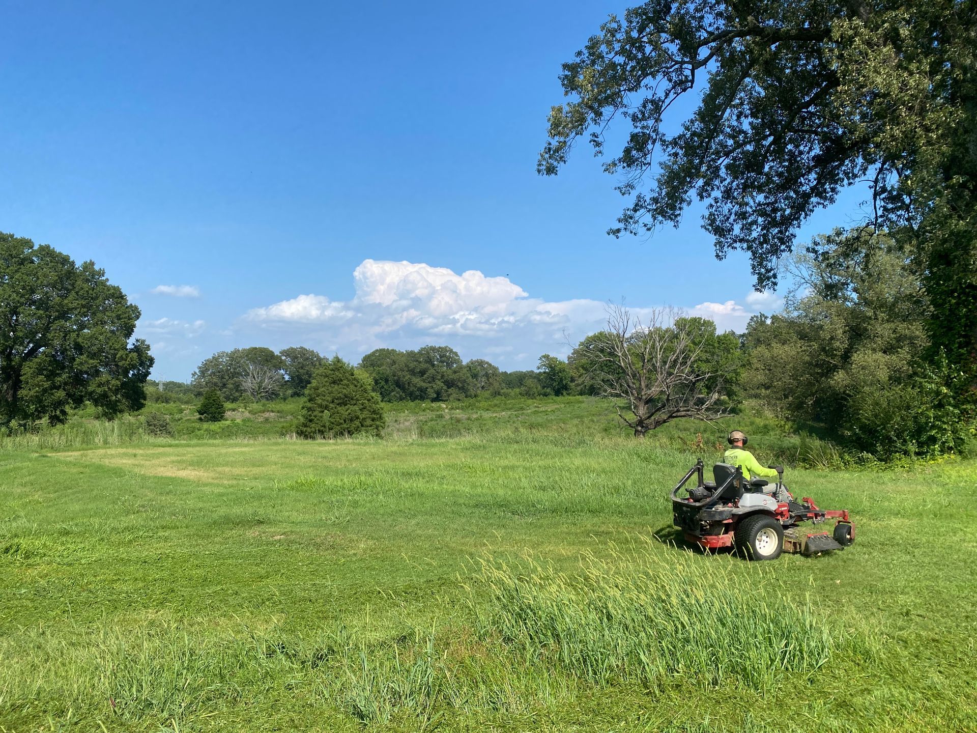 A person on a riding lawnmower cuts grass in a field on a sunny day. Trees and a partly cloudy sky are in the background.