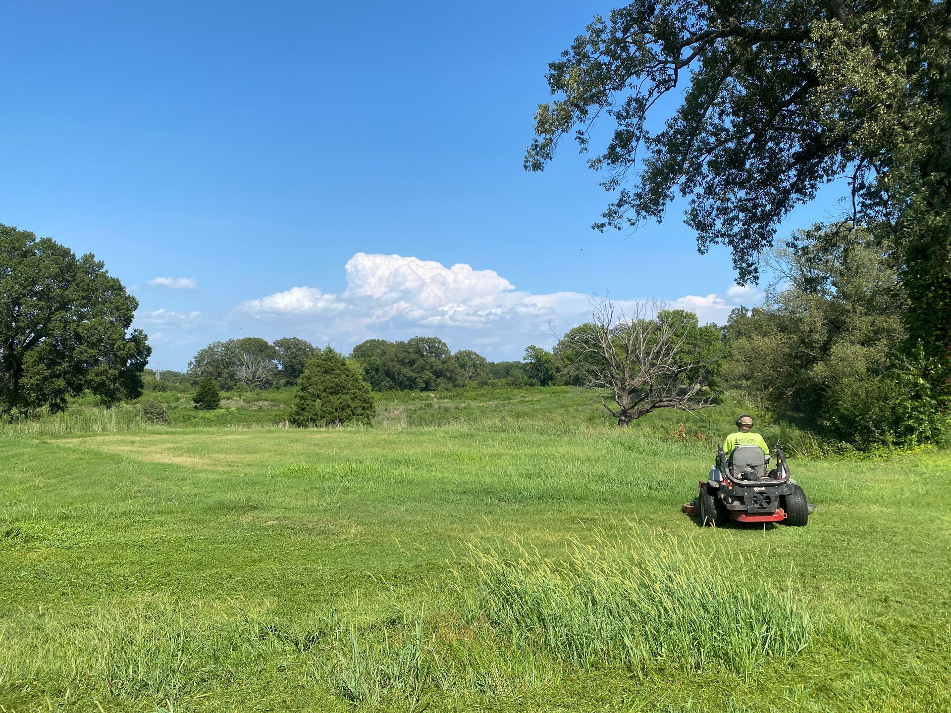 A person mows a grassy field with a riding lawn mower under a bright blue sky with fluffy clouds. Trees border the field.