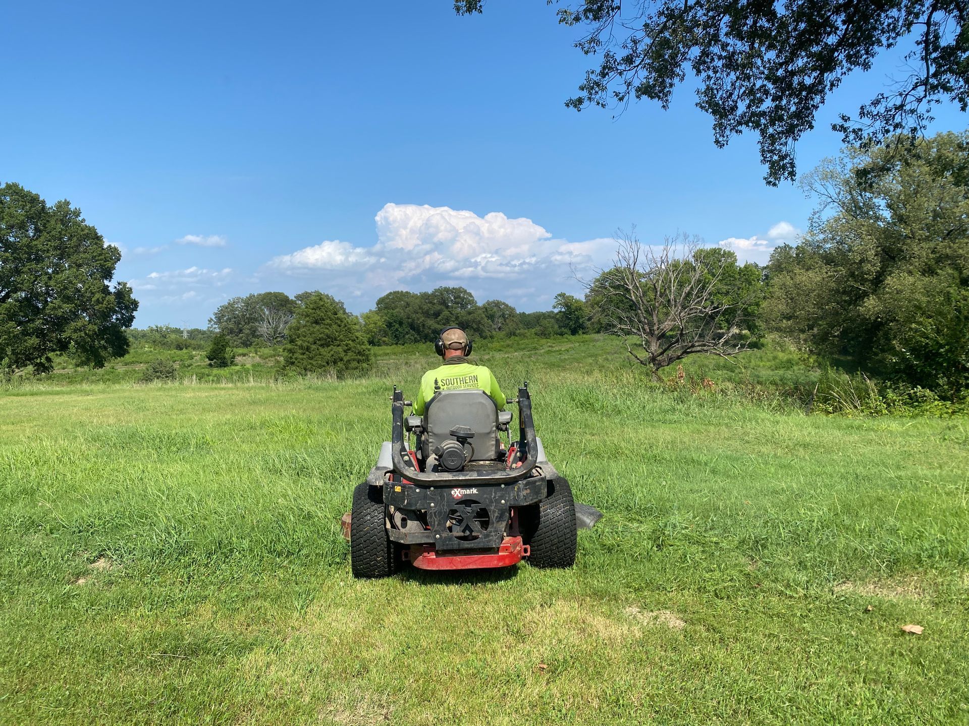 A person on a red zero-turn mower cuts grass in a field on a sunny day.