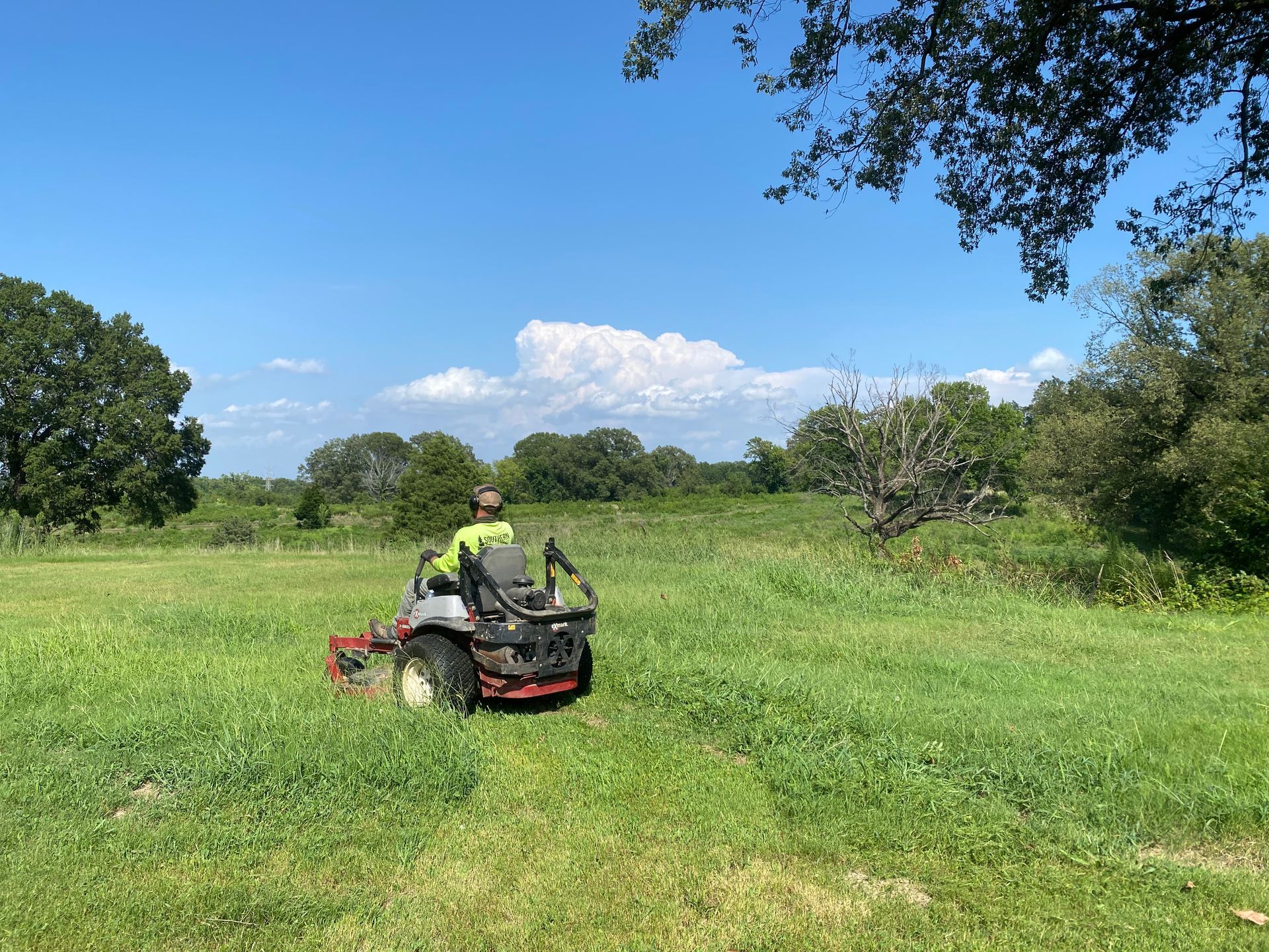 Person mowing tall green grass with a riding mower on a sunny day. Trees line the edge of the field under a blue sky.