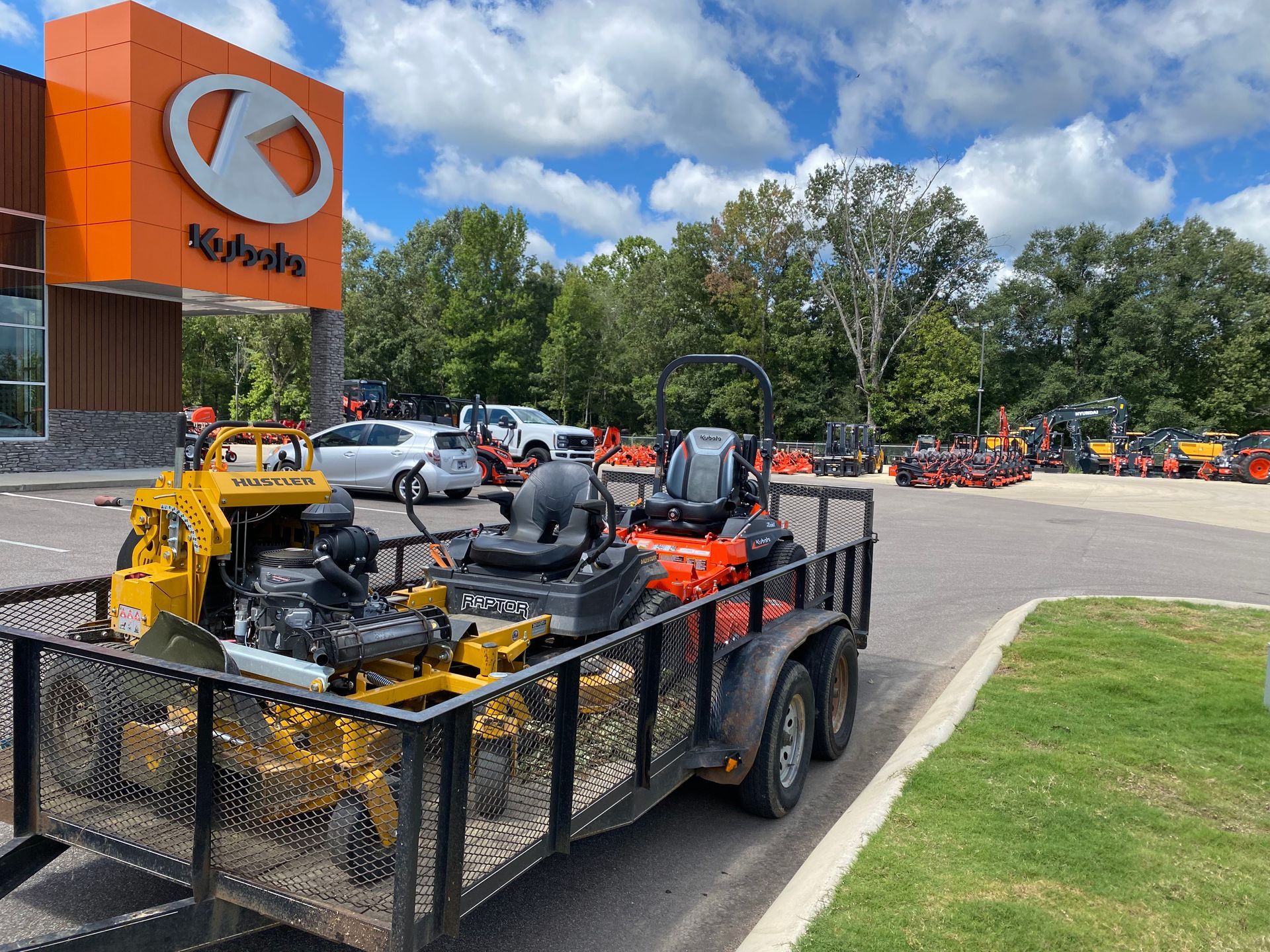 A trailer loaded with commercial lawn mowers parked in front of a Kubota dealership on a sunny day.