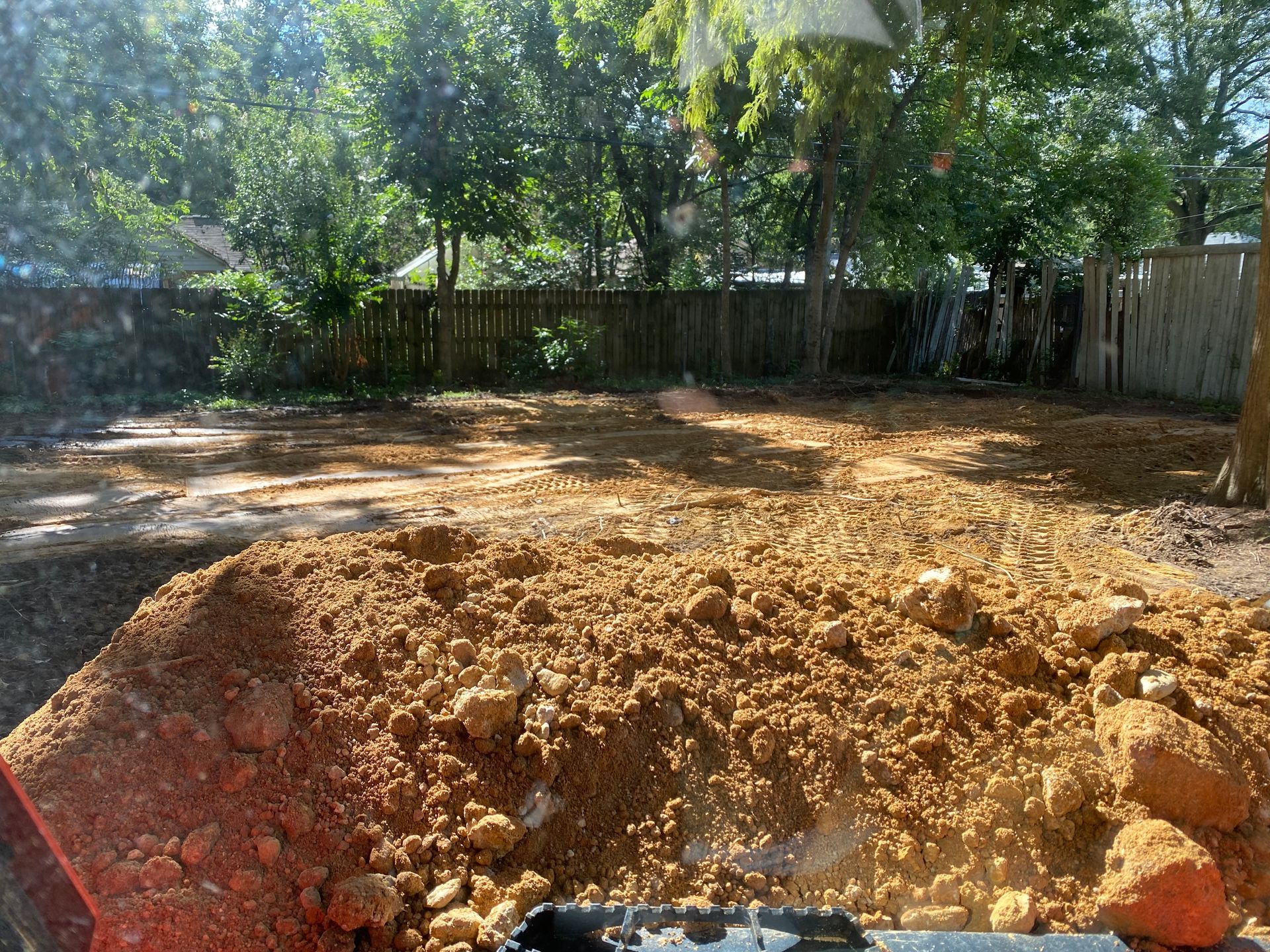 A large pile of dirt in the foreground, with a cleared backyard and a wooden fence in the background. Trees and sunlight are visible.