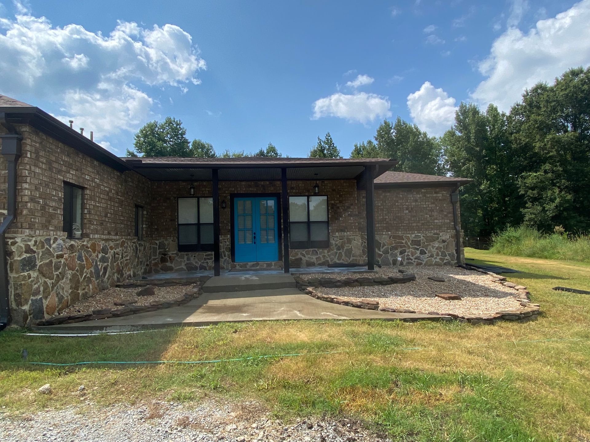 Stone-faced house with blue door, covered porch, and dark trim, set against a bright blue sky with clouds and surrounded by green grass and trees.
