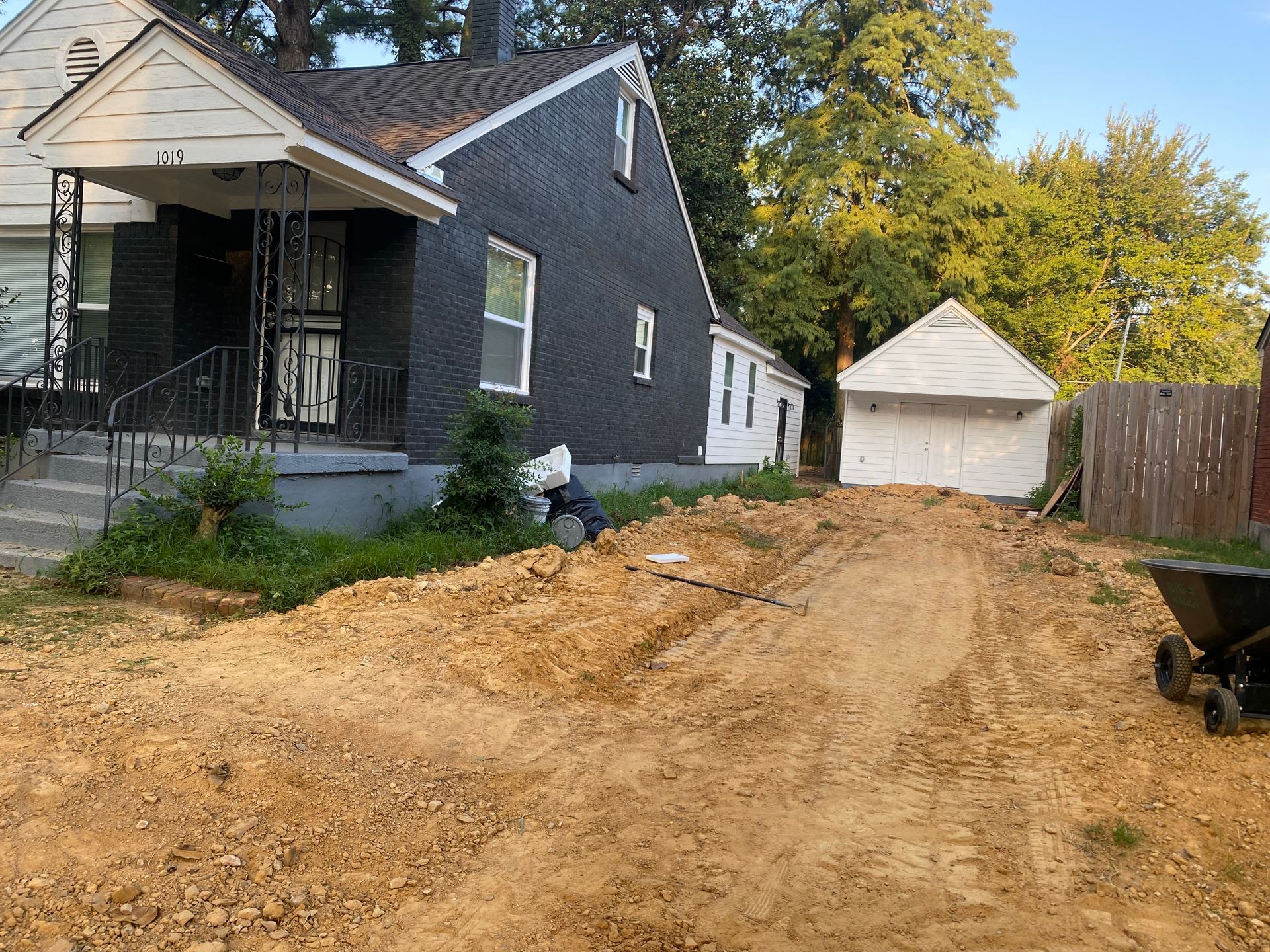 A house with black siding and a dirt driveway leading to a detached white garage.