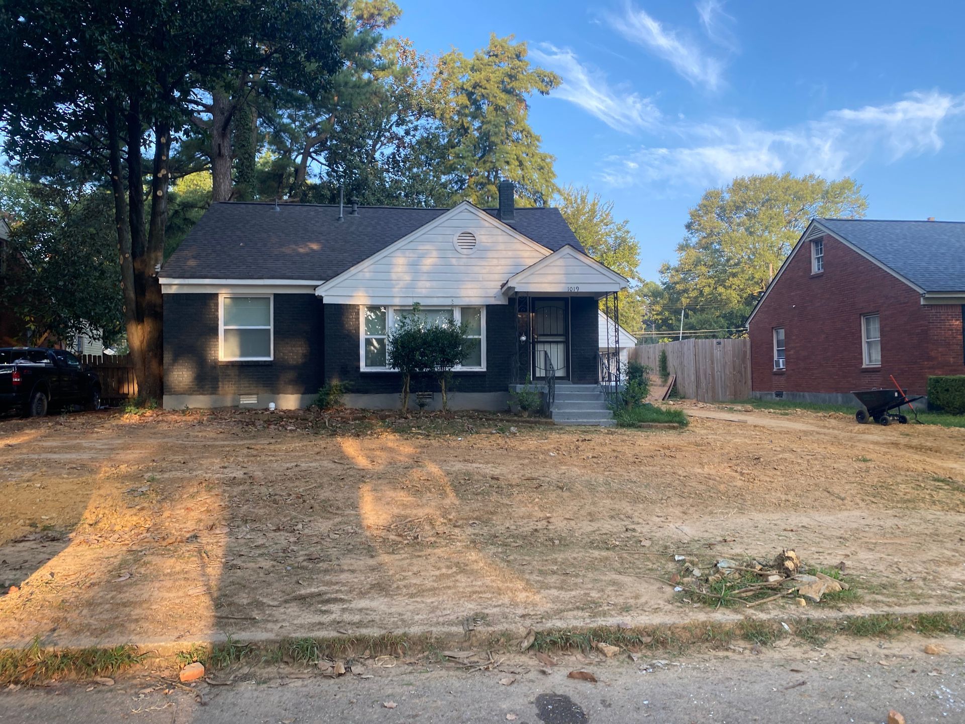 A dark gray house with a brown roof and a dry, brown front yard, next to a red brick house and trees.