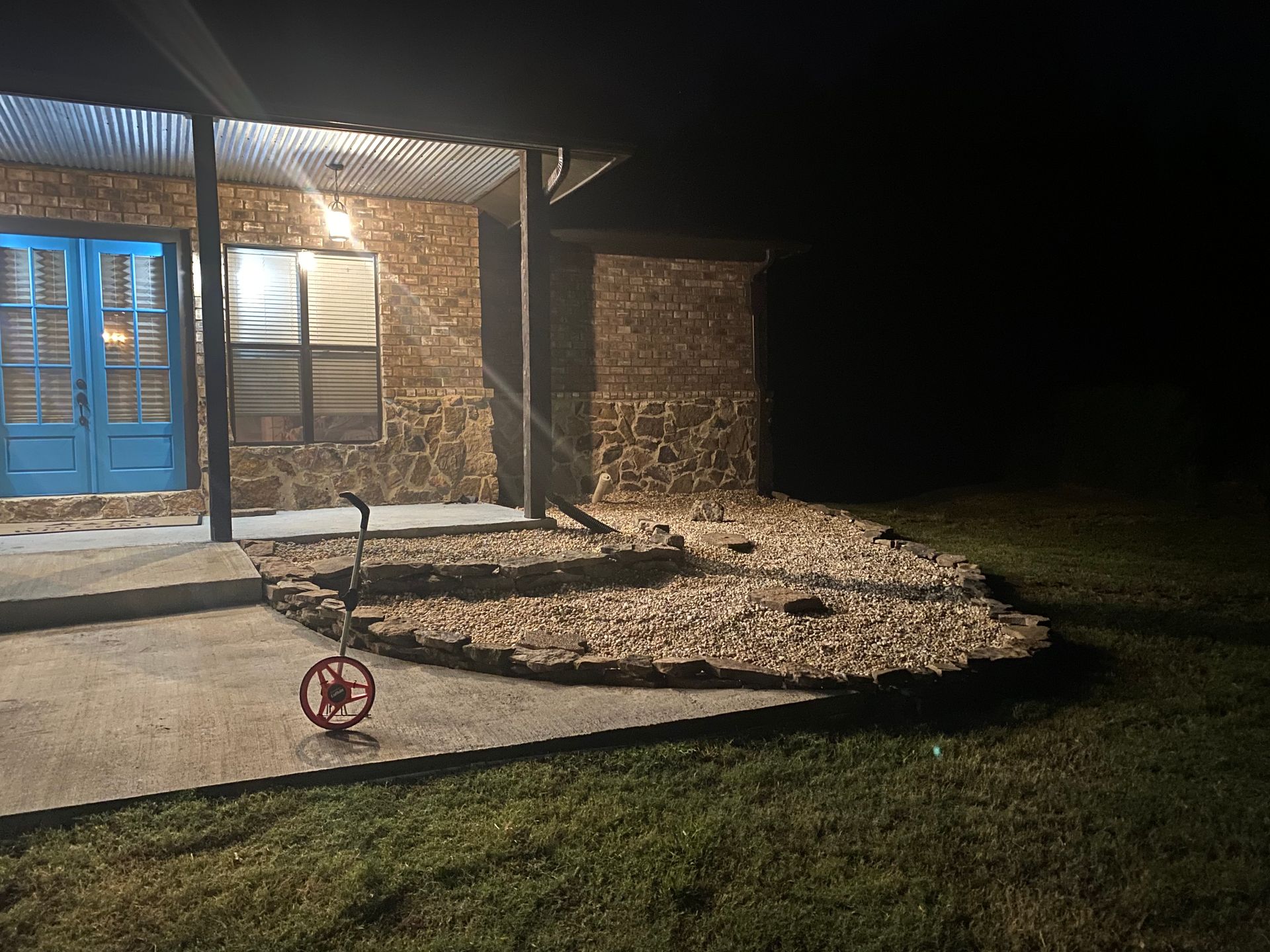 A stone house exterior at night with blue doors and a covered porch. Landscaping with rocks in front.