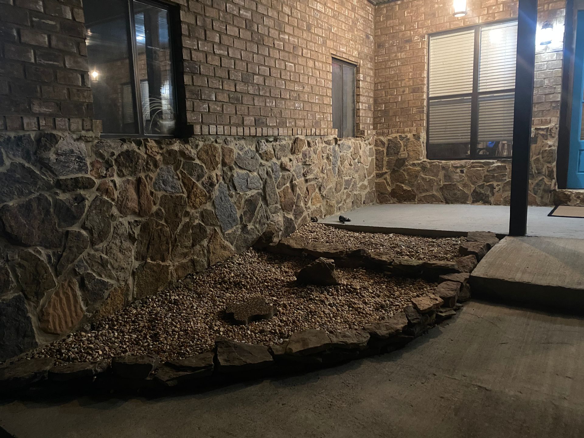 Exterior view of a building with a stone foundation, gravel landscaping, and a brick upper level. A door and window are visible.
