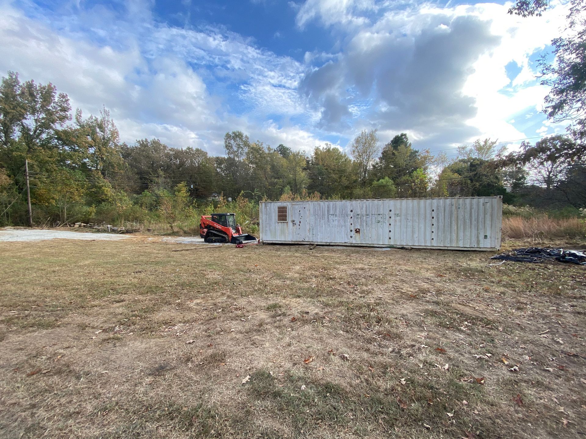 A small orange tractor next to a weathered white shipping container in a field with trees in the background under a cloudy blue sky.
