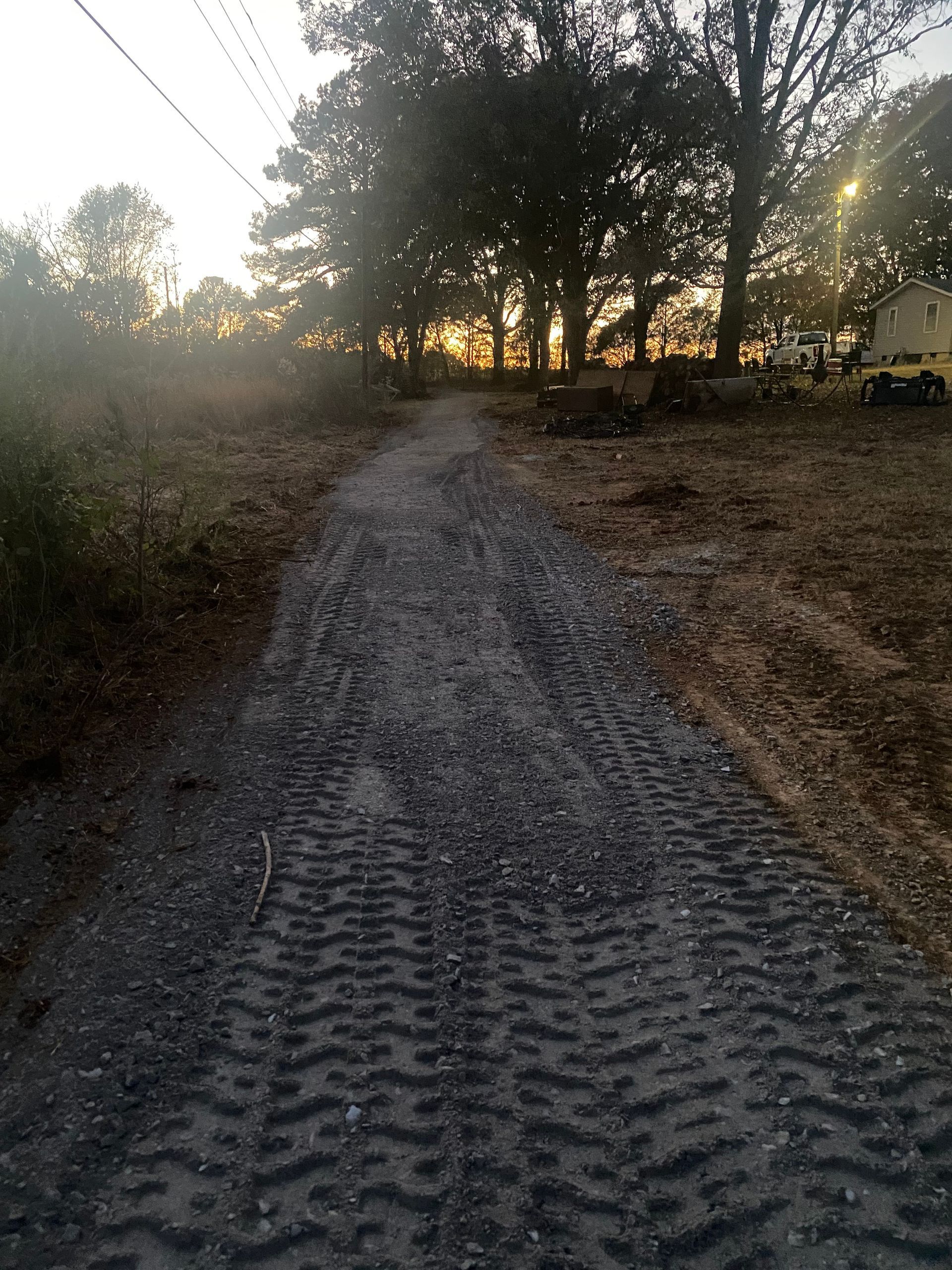 Gravel driveway leading towards a treeline at sunset, with tire tracks visible. Yellow vehicle and trees are in the background.
