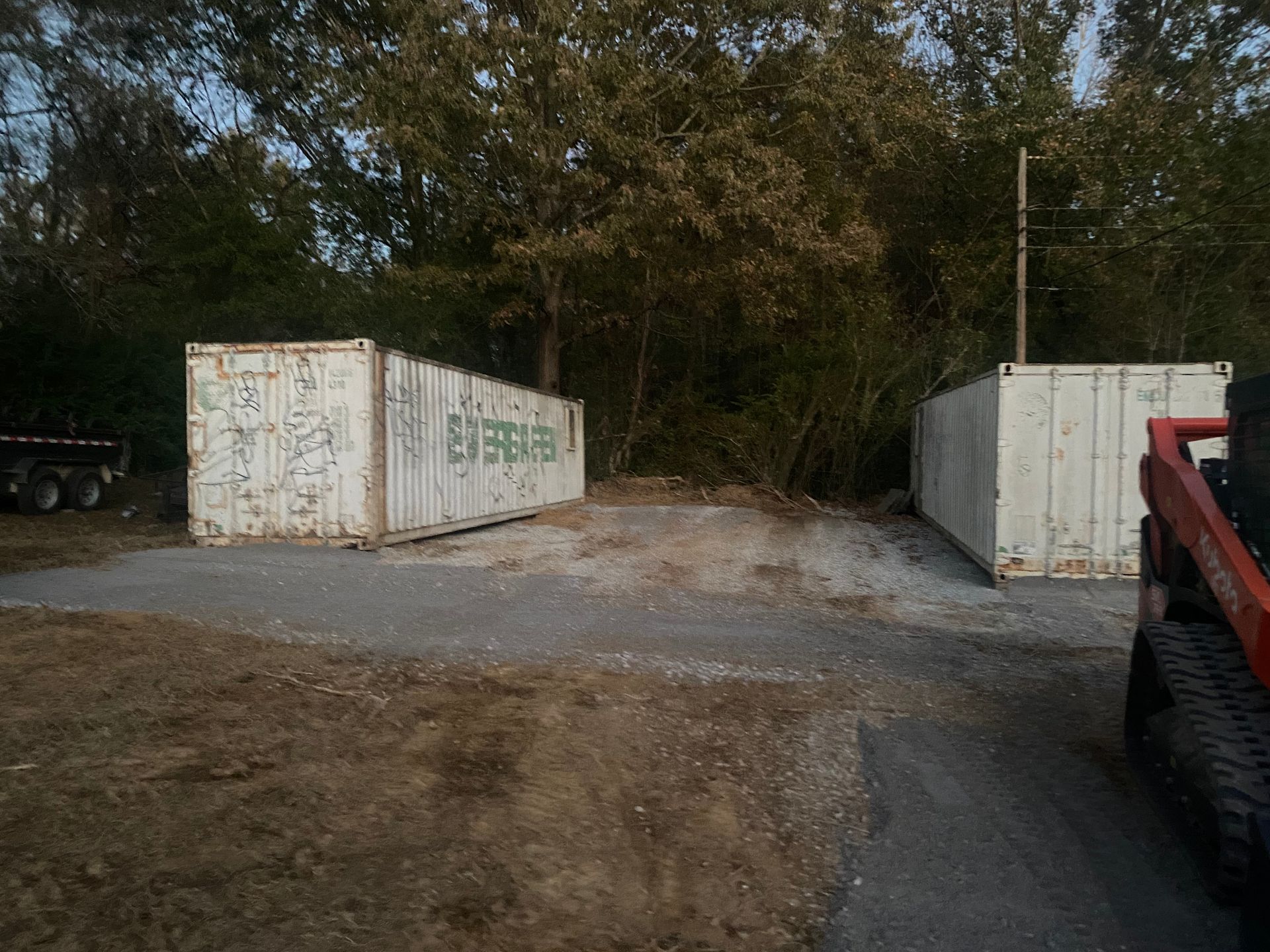 Two white shipping containers sit on gravel, with a skid steer on the right. Trees and a trailer are visible in the background.