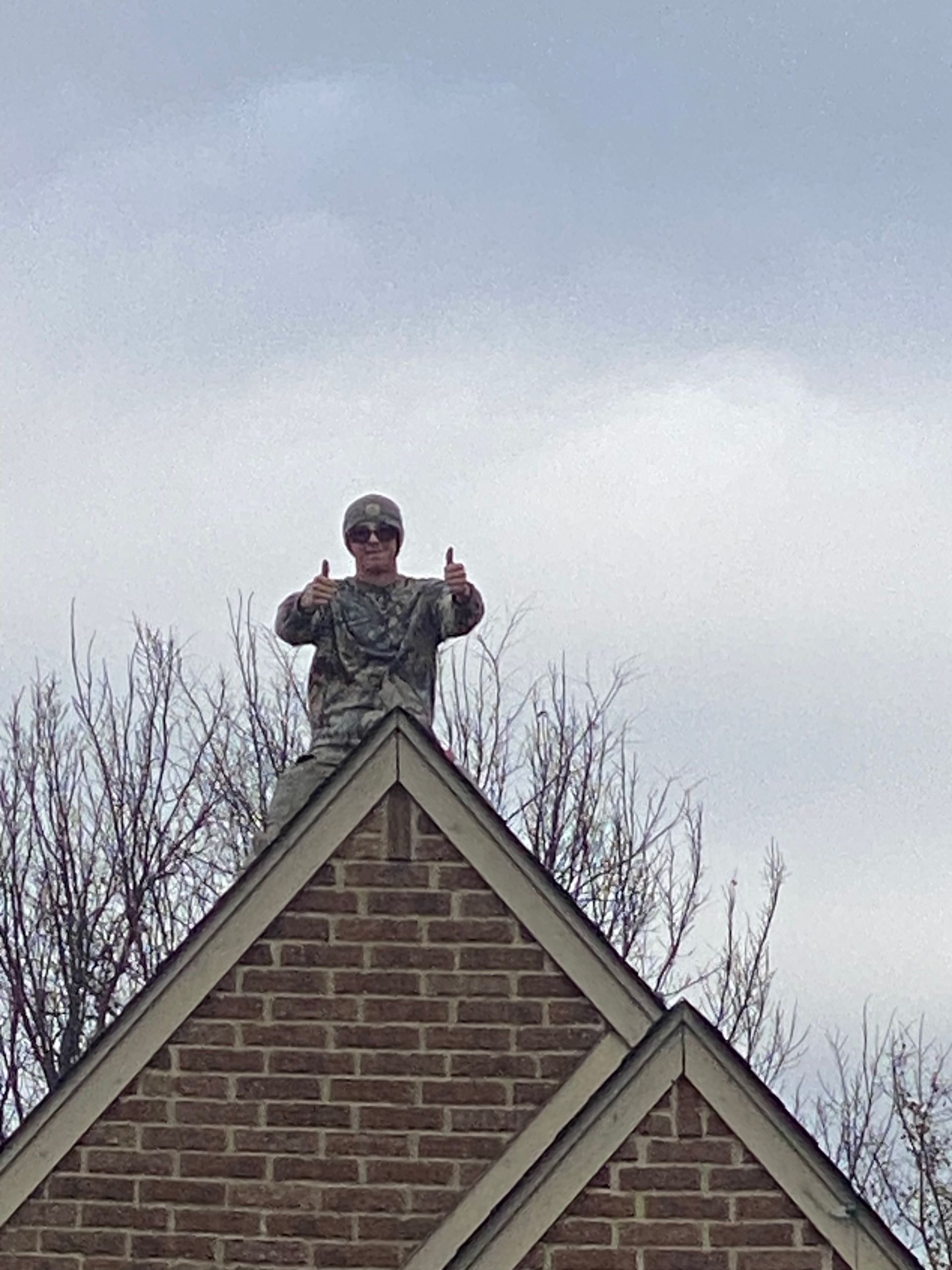 Person in camouflage uniform on a brick-covered rooftop, giving thumbs up. Gray sky in the background.