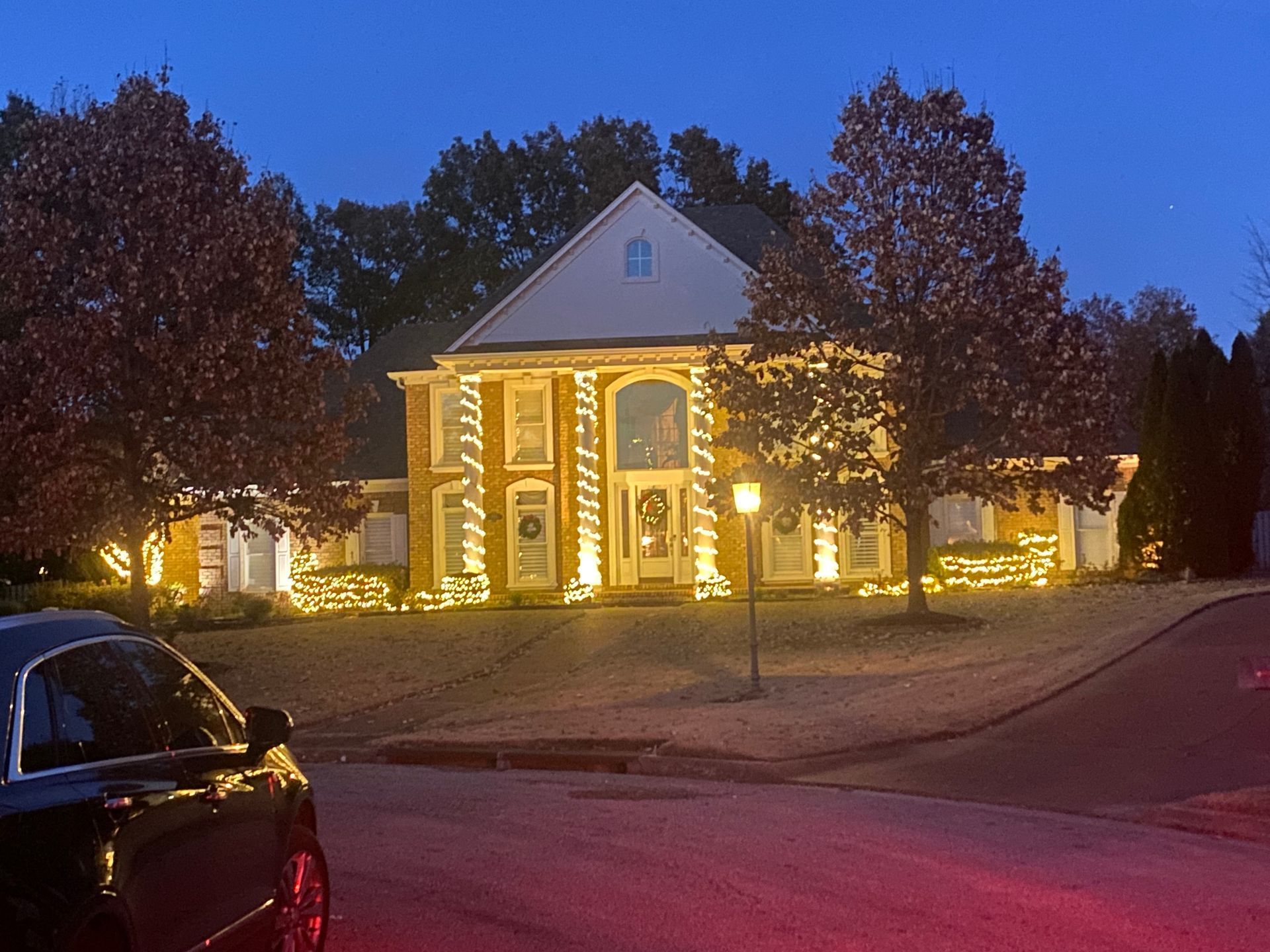 A two-story brick house decorated with warm white Christmas lights at dusk. A car is parked in the foreground.