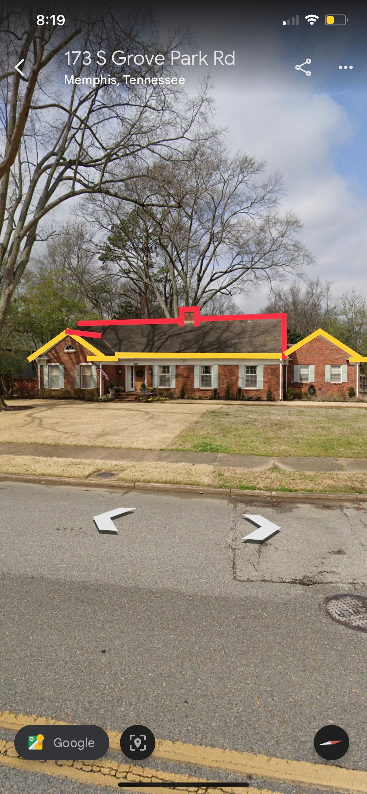 A brick house with yellow trim and a red roof on a tree-lined street. The image is from a street view.