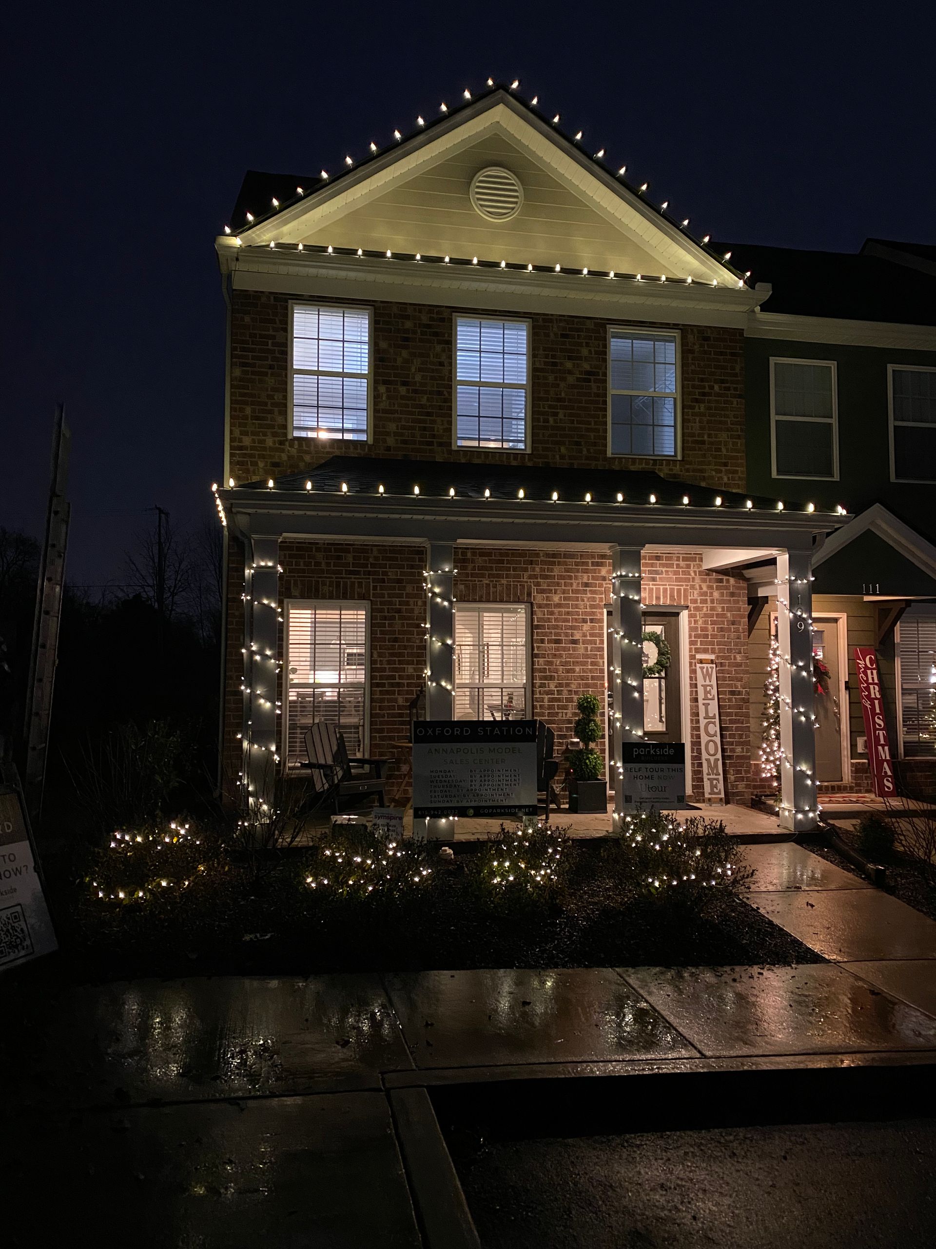 A brick townhome is decorated with white Christmas lights. Lights outline the roof, porch, and bushes in front. It's nighttime.
