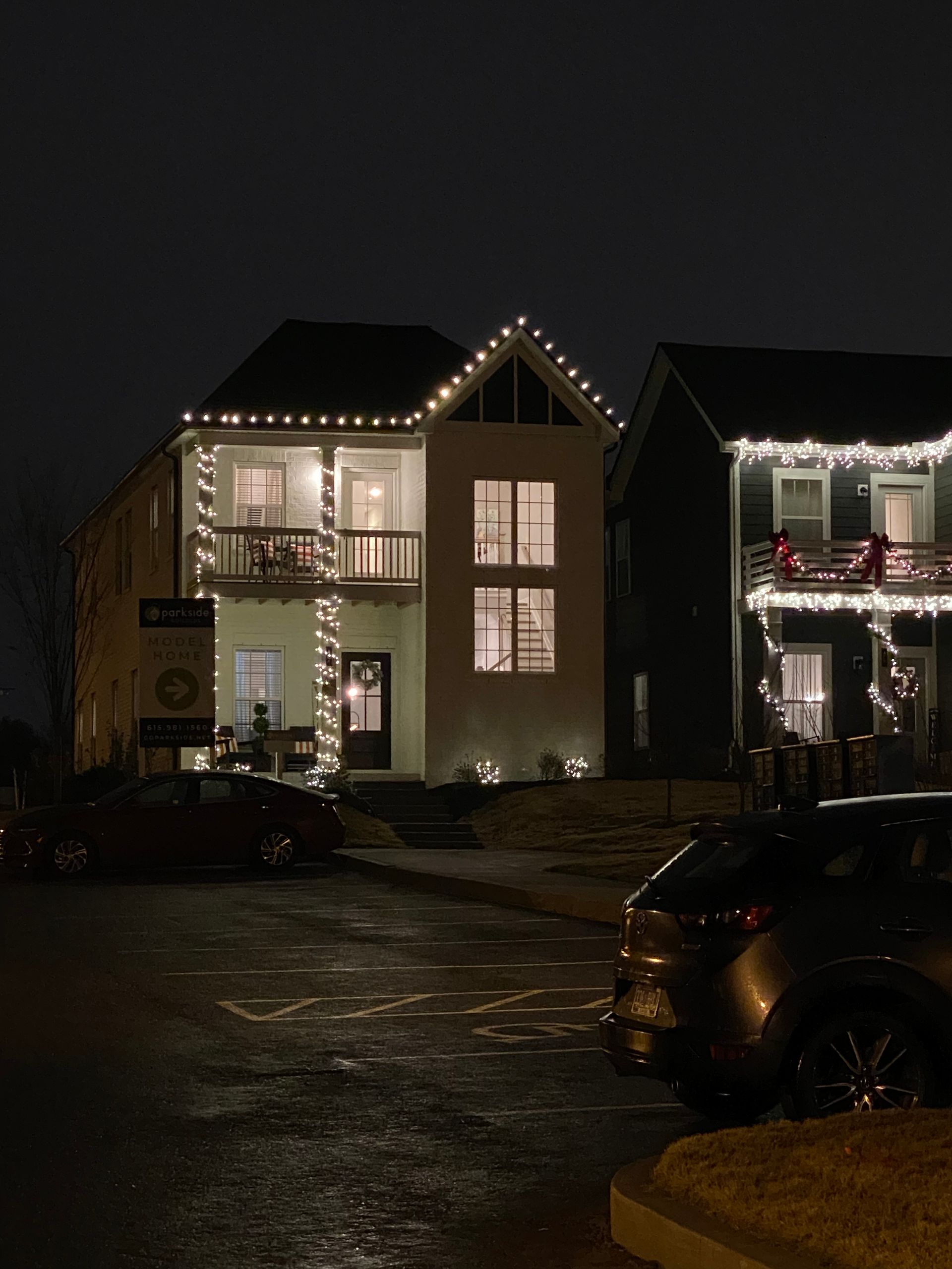 Two-story house and adjacent house decorated with Christmas lights at night. Cars parked on street.