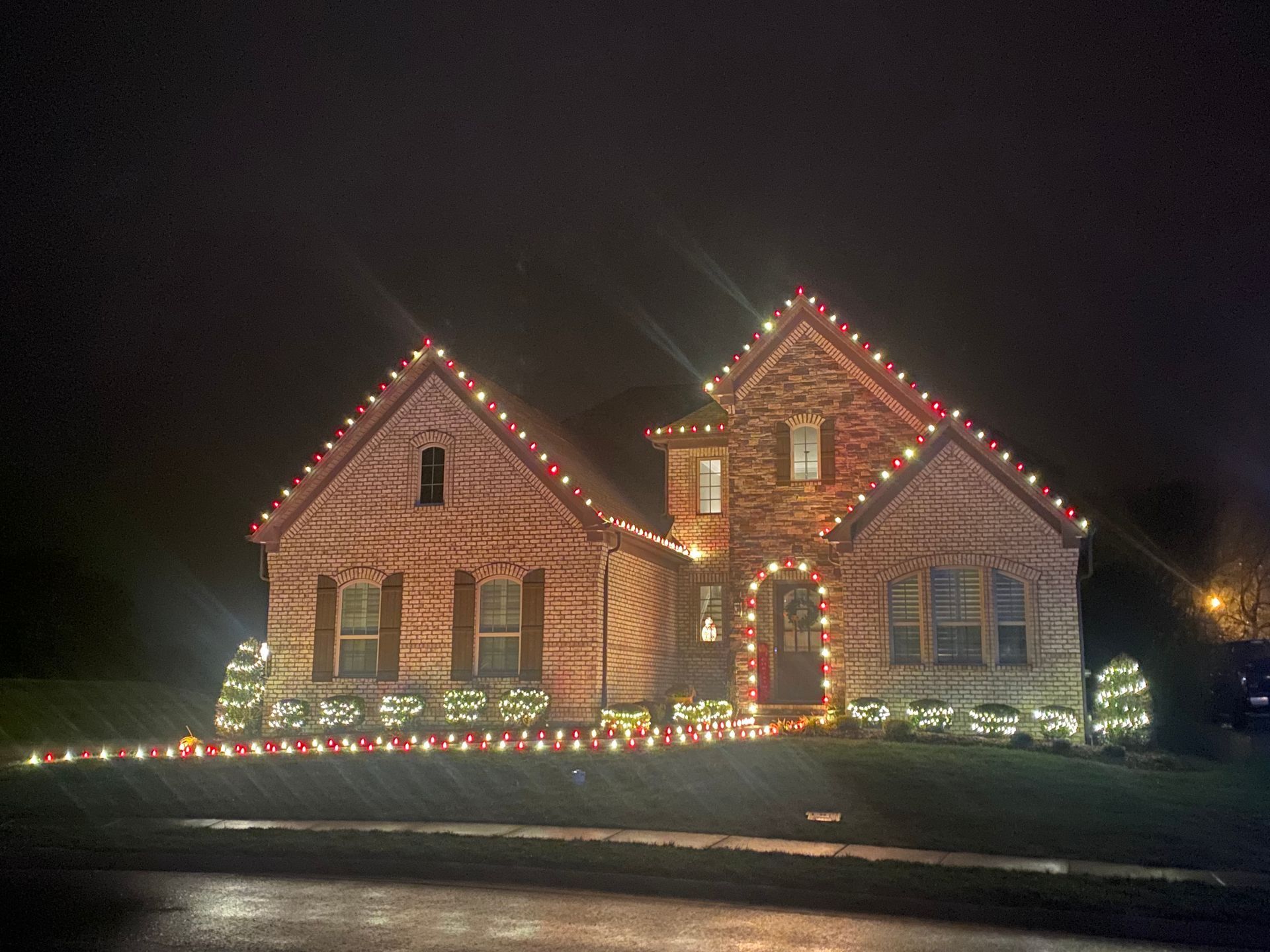 A brick house is decorated with red and white Christmas lights. Lights frame the roofline, windows, and bushes.