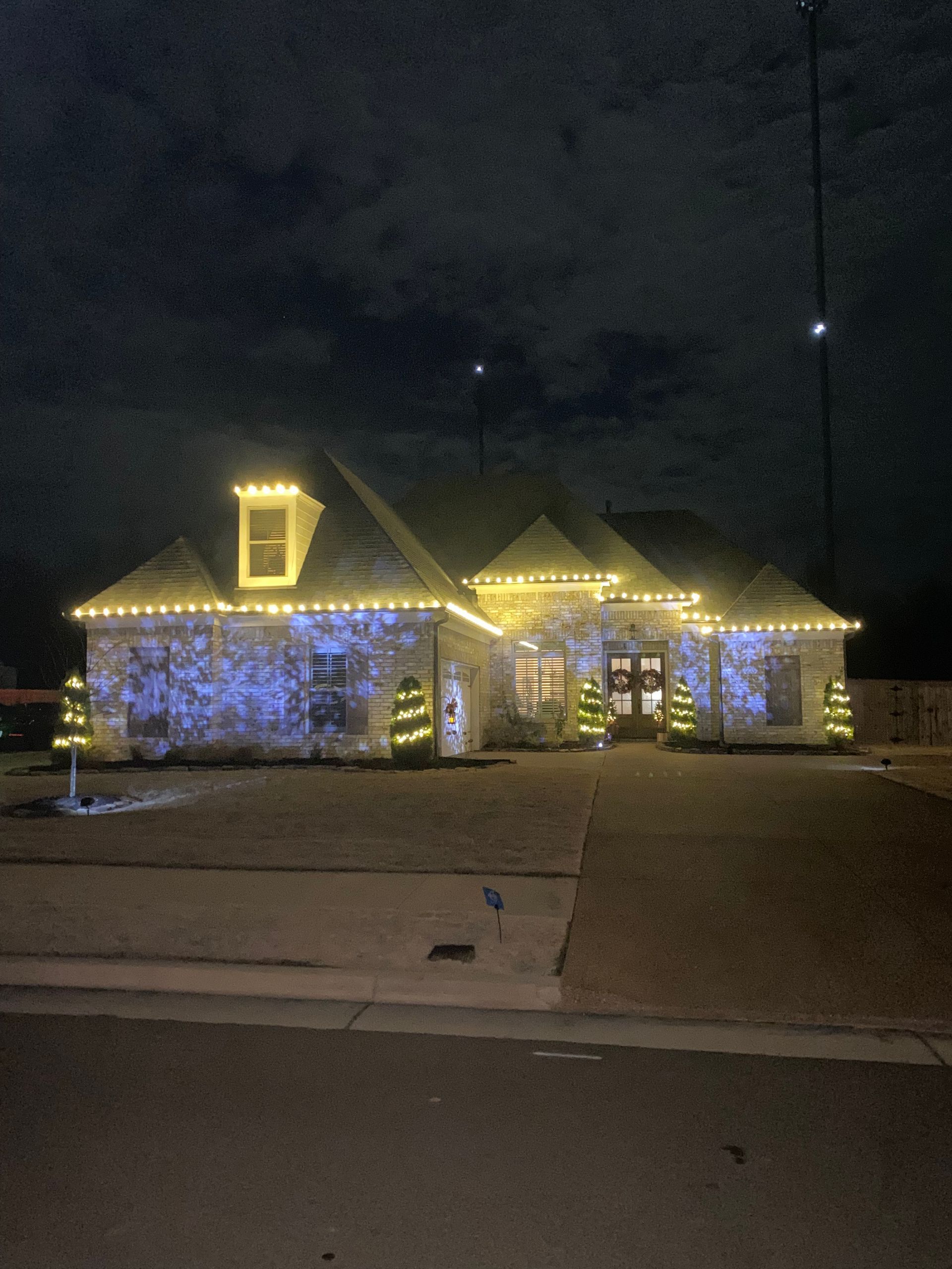 A house at night, decorated with yellow Christmas lights outlining the roof and blue lights on the stone exterior. Small lit Christmas trees flank the front door.