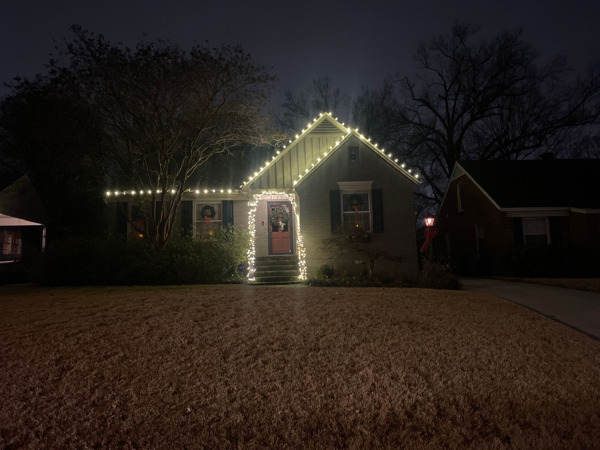 A house at night, lit with white Christmas lights outlining the roof and doorway, set against a dark sky.