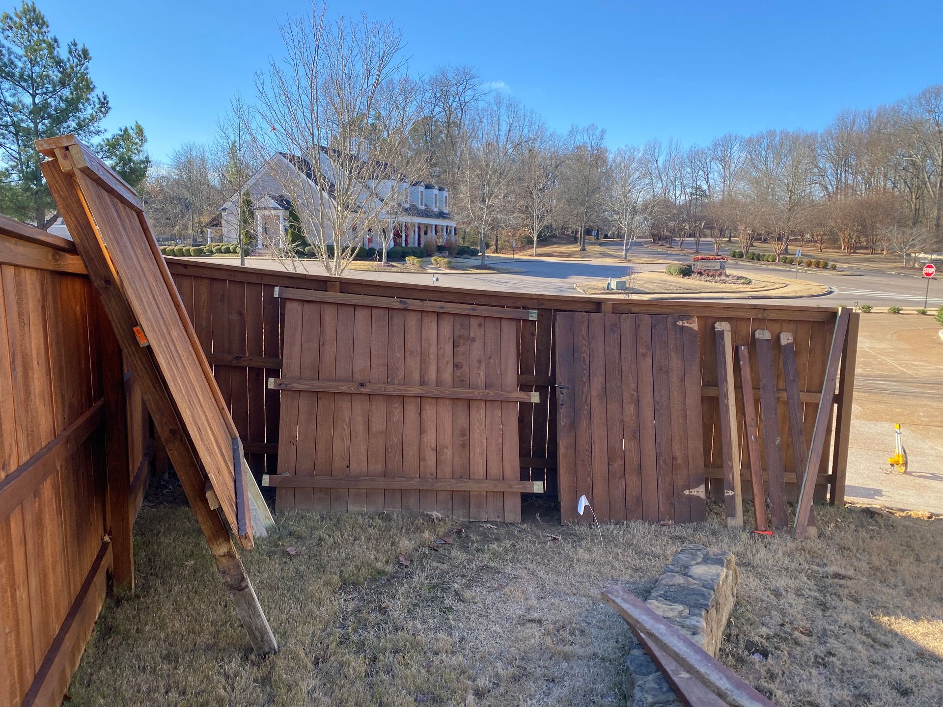 Damaged wooden fence in a grassy yard with a house and trees in the background under a blue sky.