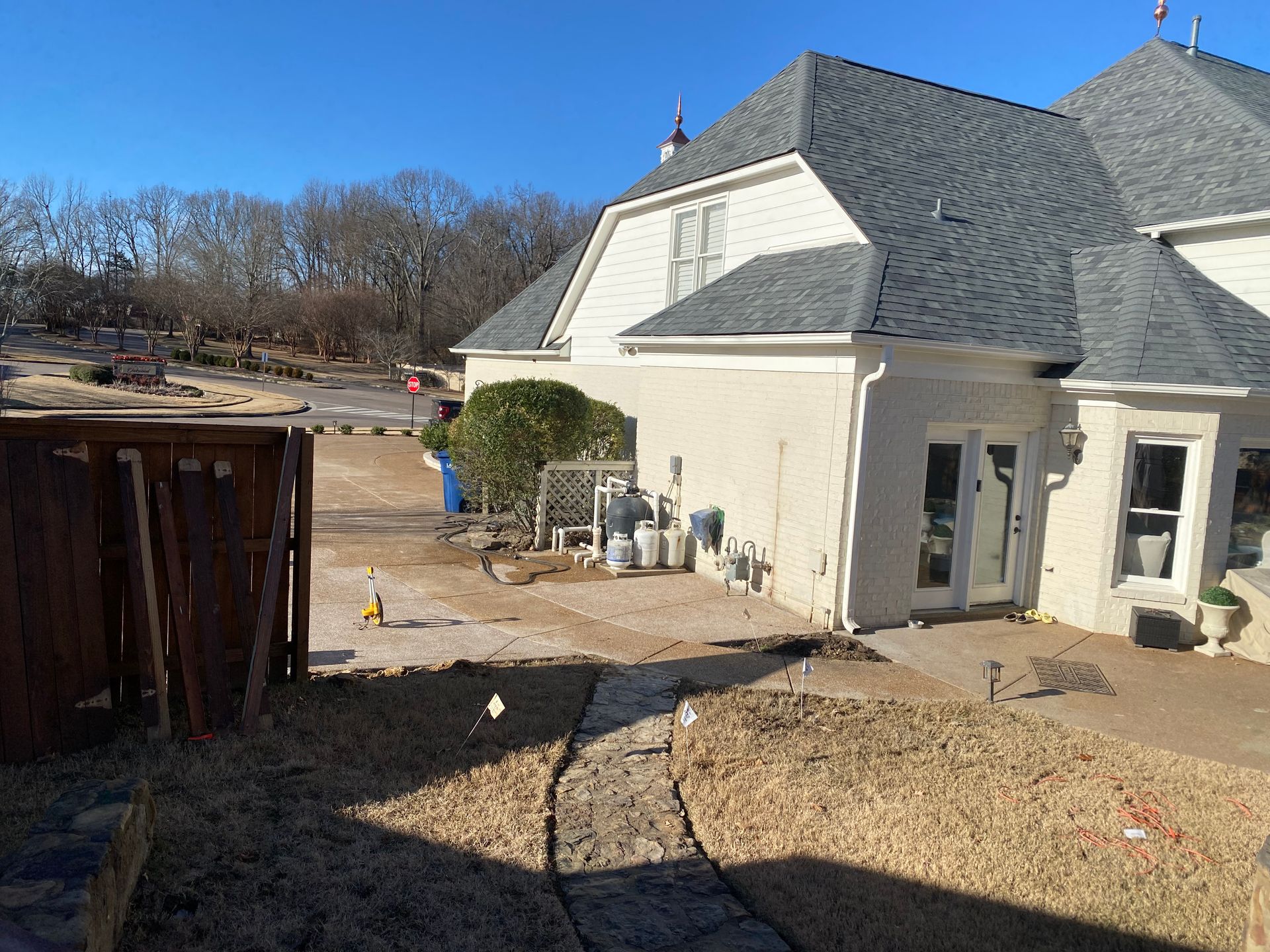 A house with a gray roof and white siding, a walkway, and brown fence. A road and trees are in the background.