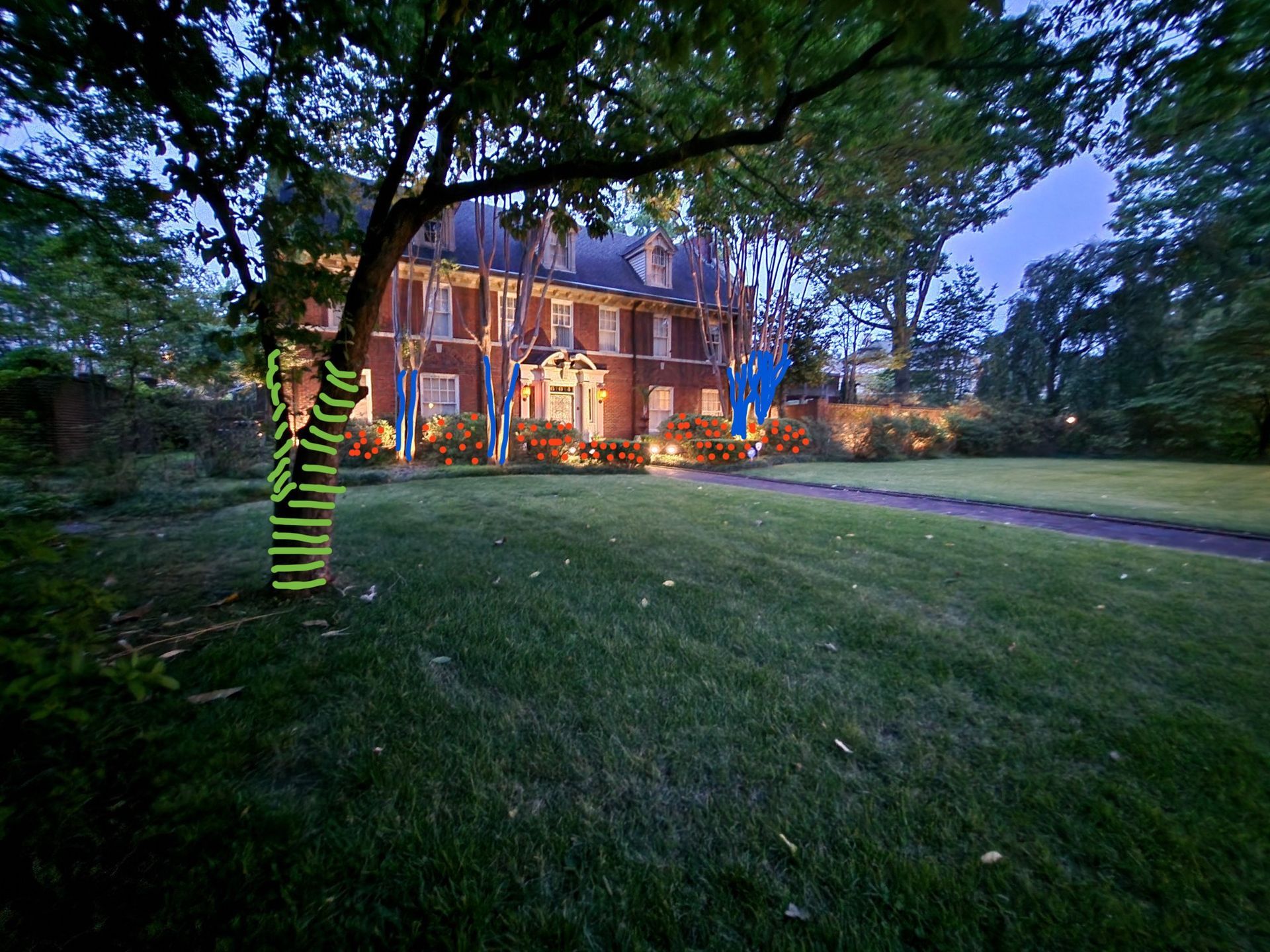 A large brick house at dusk with a wrapped tree and illuminated lawn. Green and yellow tape decorates the tree trunk.