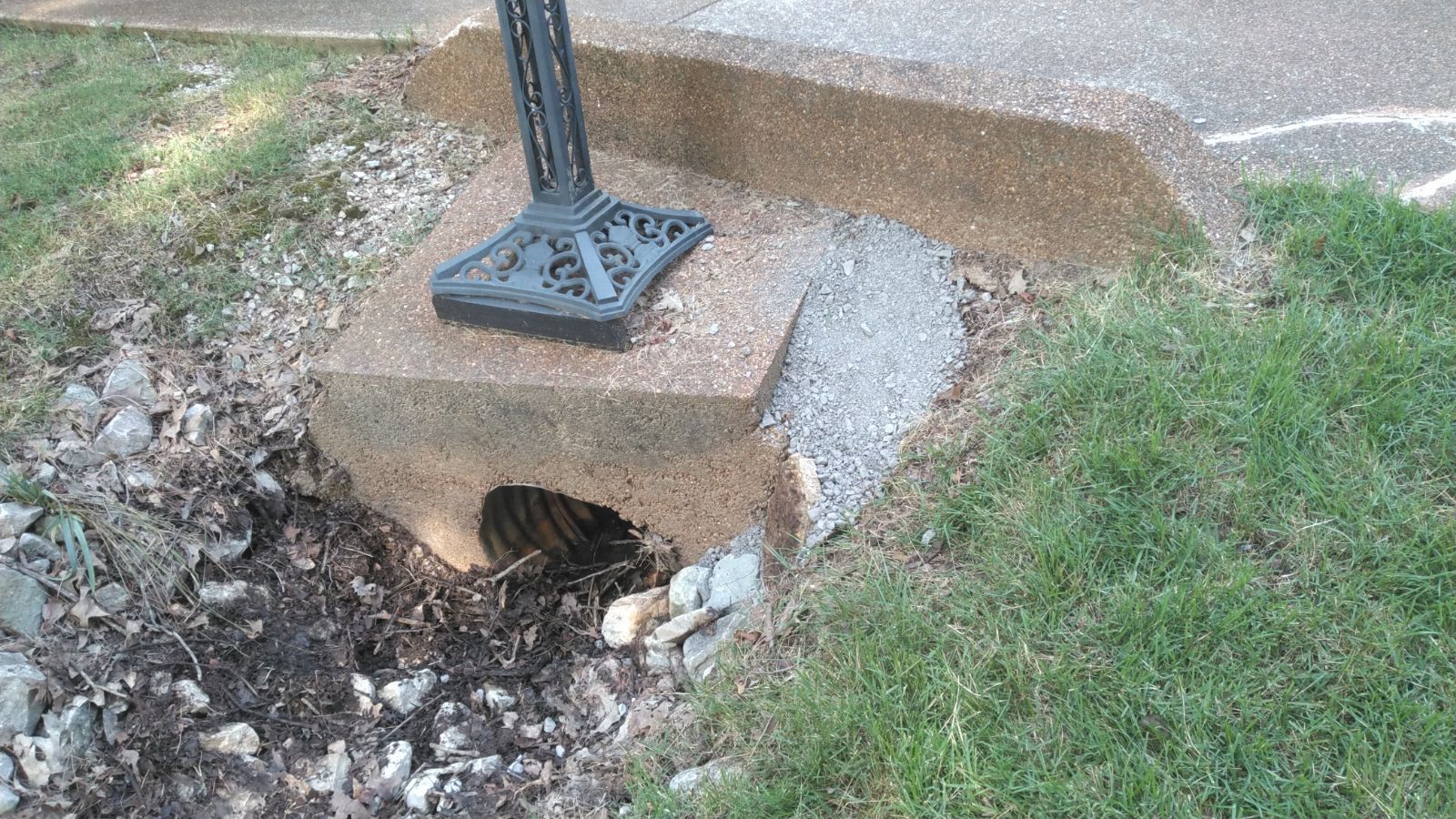 Concrete drain with a metal grate and a black lamp post section, set in grass beside a road.