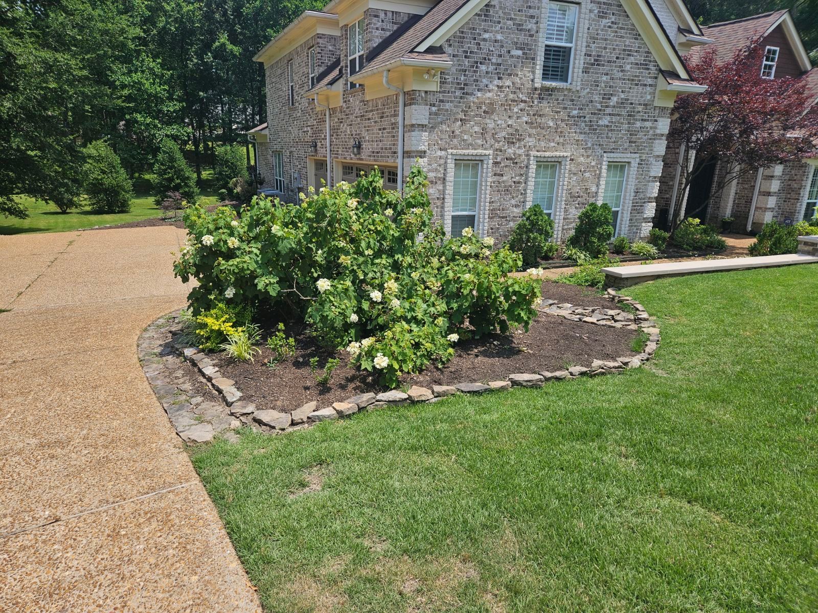 Stone house with a circular flowerbed in front, filled with green bushes and white flowers. A gravel driveway and green lawn are also visible.