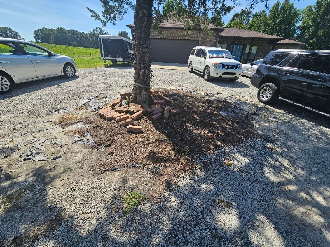 Gravel driveway with a tree at its center, surrounded by brick. Cars and a trailer are parked around the tree.