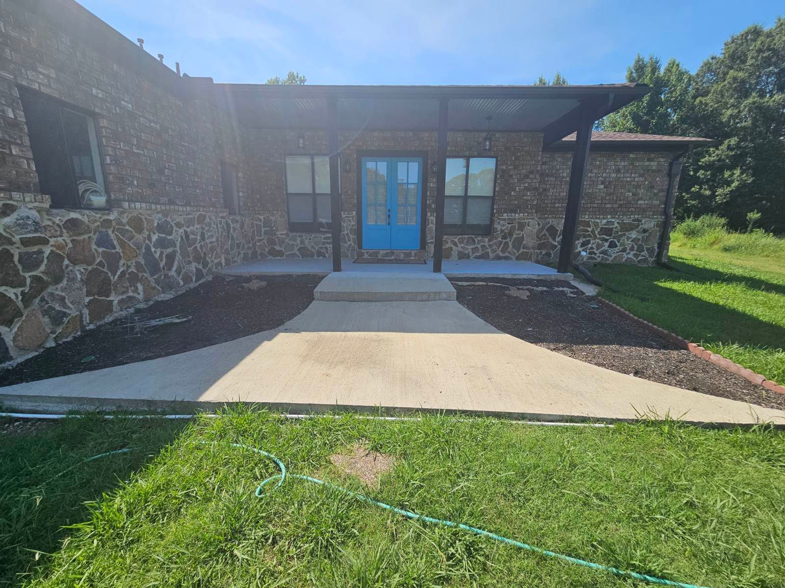 A blue front door of a stone house with a concrete walkway and dark mulch beds in a sunny yard.