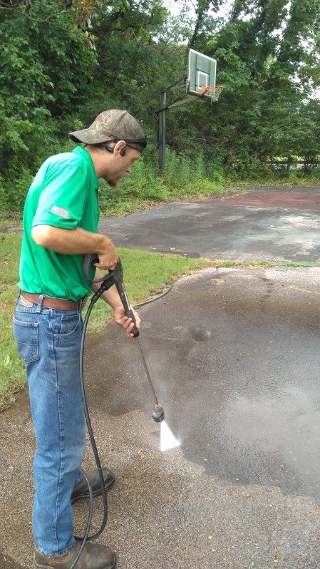 A man in a green shirt and baseball cap pressure washes a concrete surface. He stands outside near a basketball hoop.
