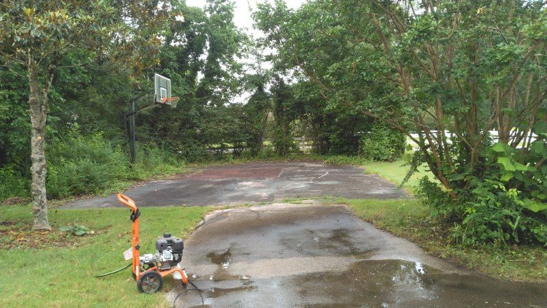 A pressure washer sits on a wet driveway, cleaning a concrete basketball court in a yard surrounded by trees.