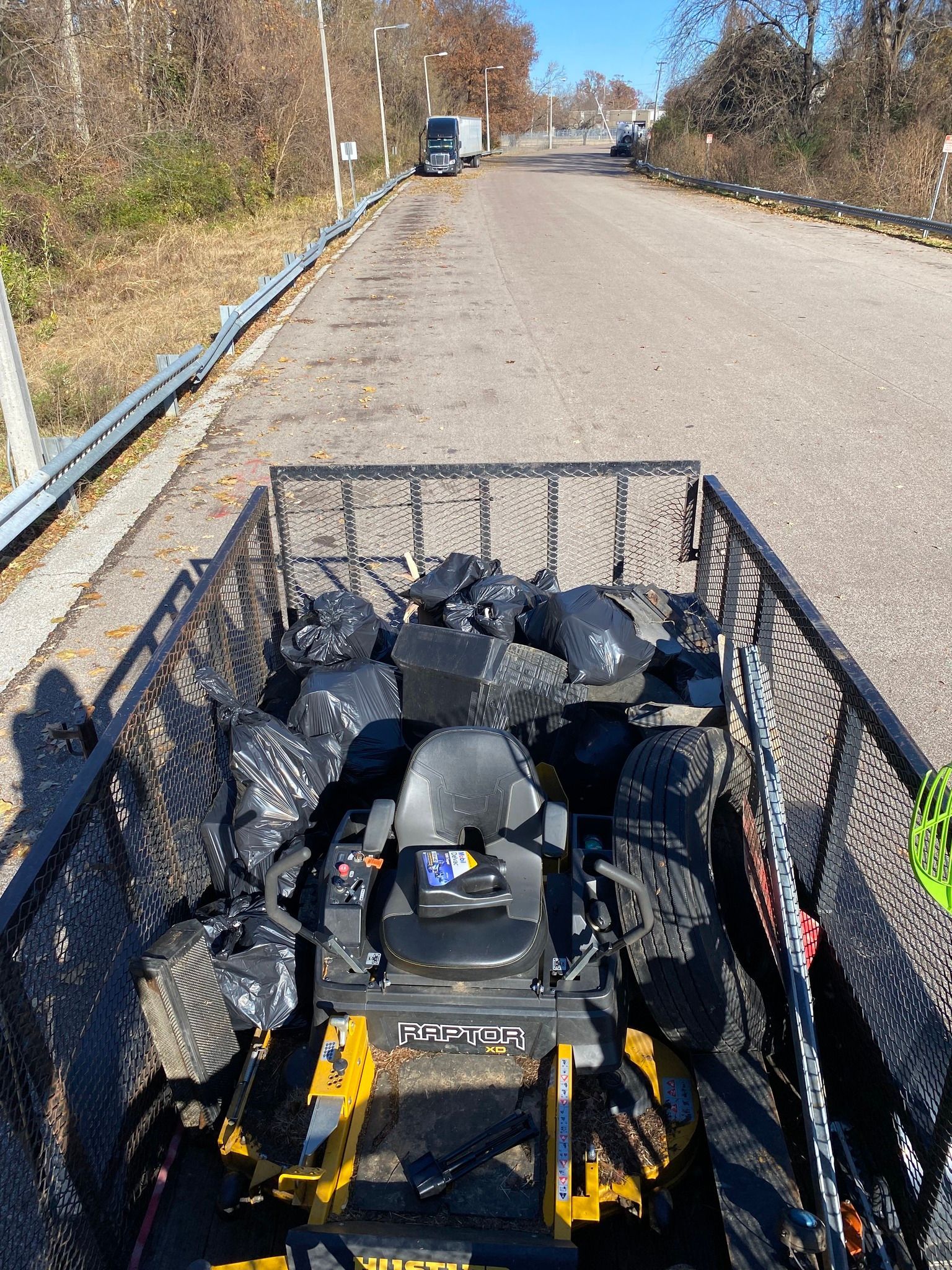 A trailer filled with black trash bags and a riding lawnmower on a road.