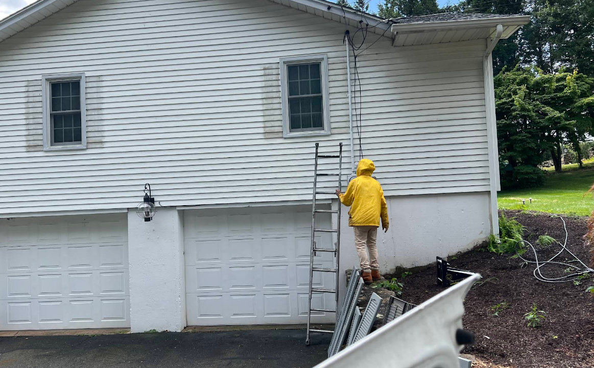 A man in a yellow jacket is standing on a ladder in front of a white house.