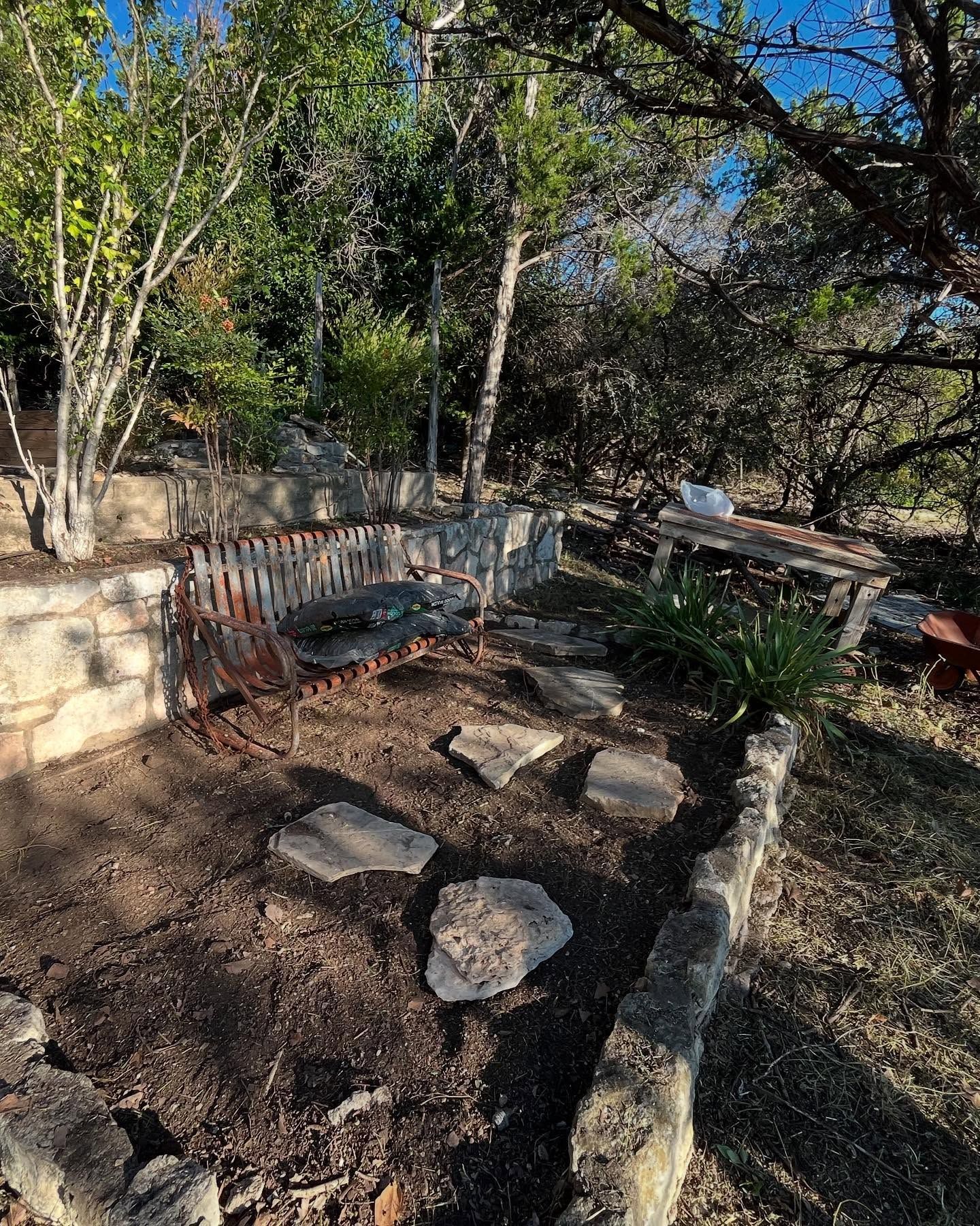 A garden with a rustic bench, stone pathway, and retaining walls, surrounded by trees under a blue sky.
