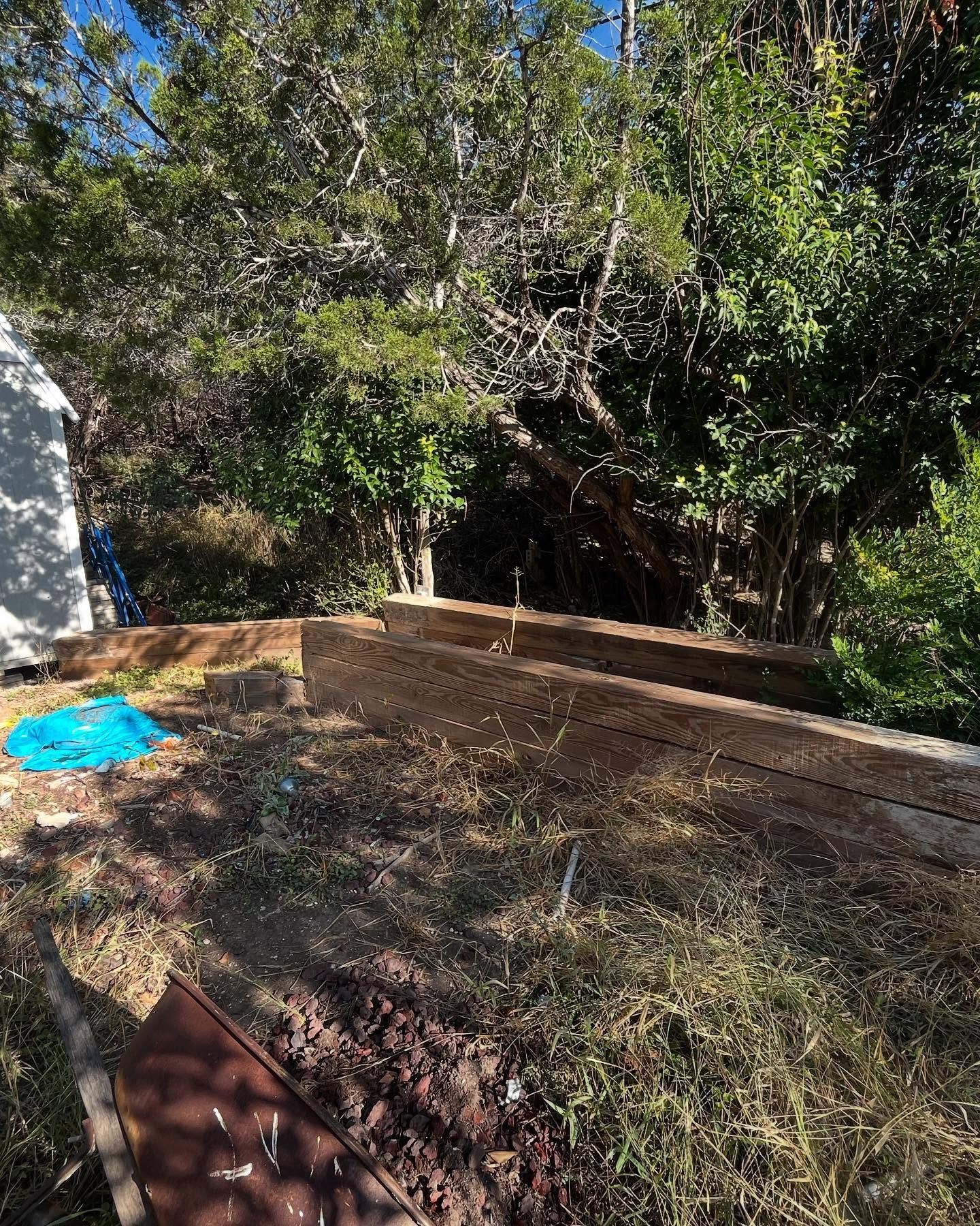 Raised garden beds with soil, next to a wooded area and a structure on the left.