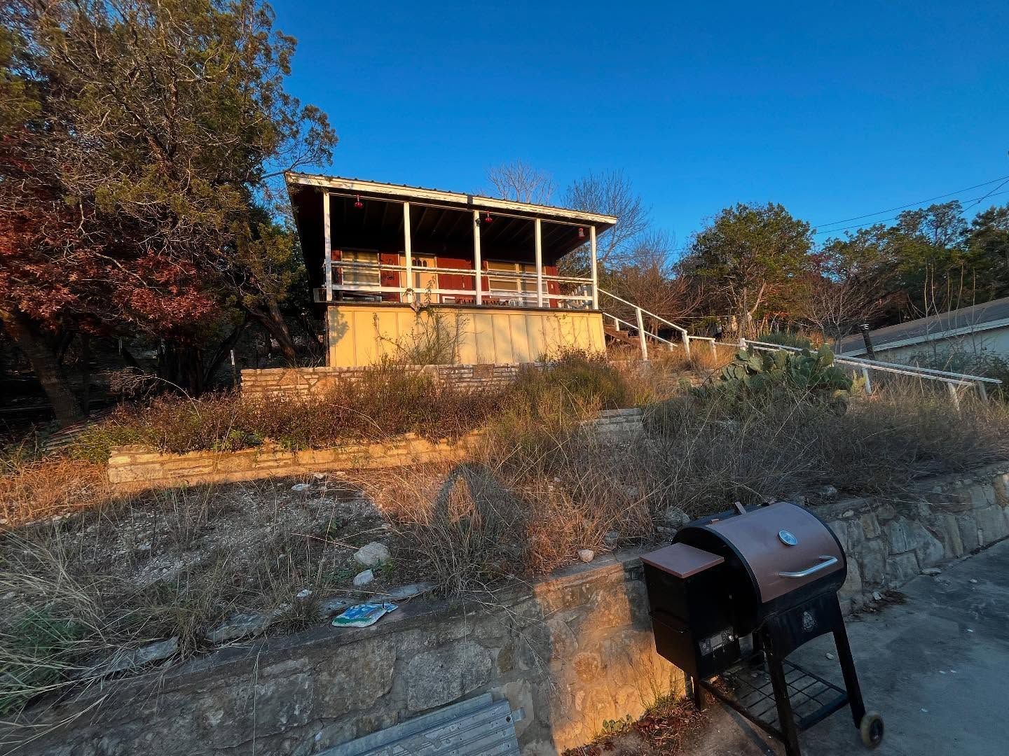 Small, weathered cabin with a porch on a hillside with a smoker grill in the foreground. Blue sky.