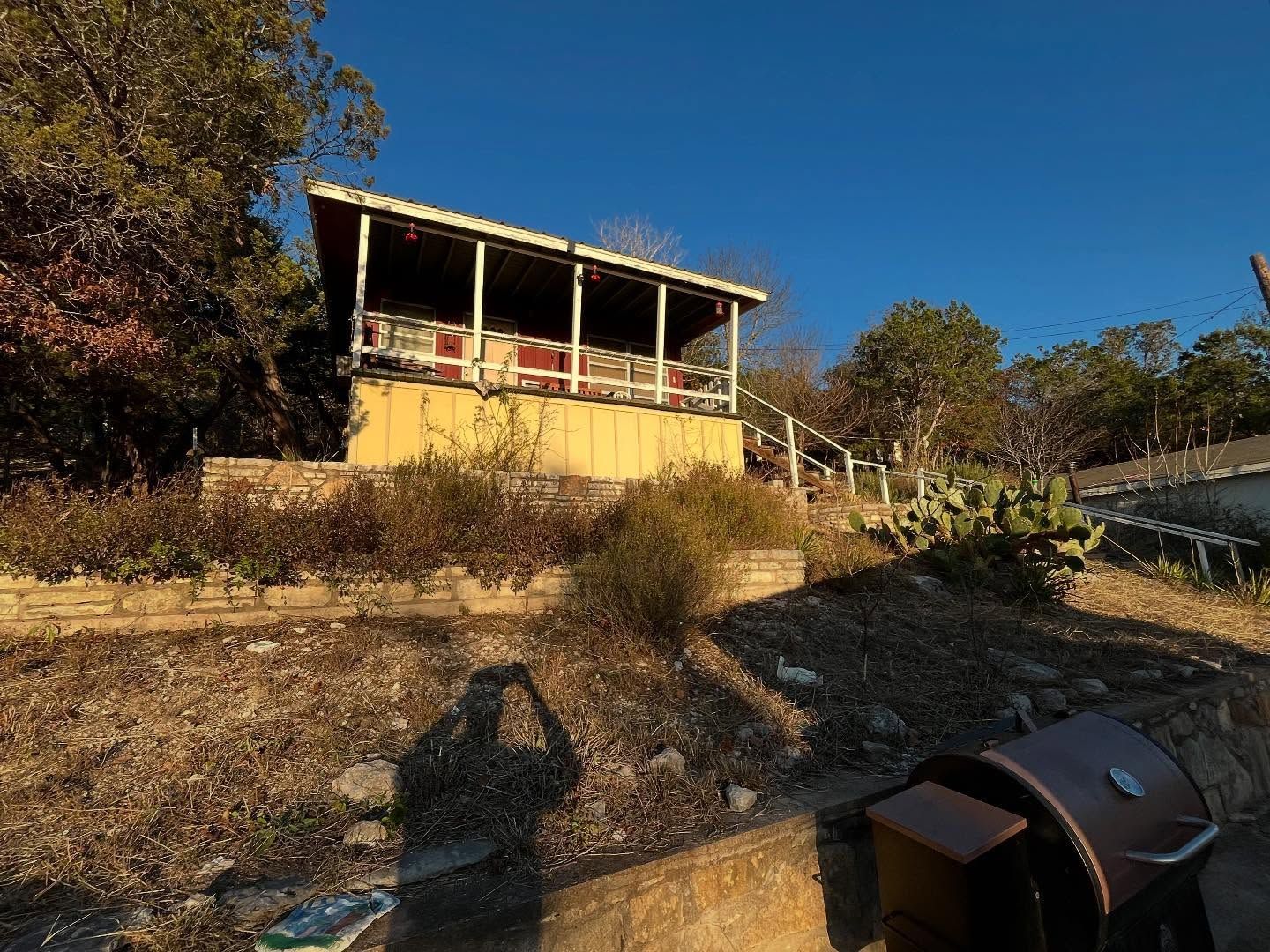 Yellow cabin on a hillside with a porch, surrounded by dry brush and a blue sky.