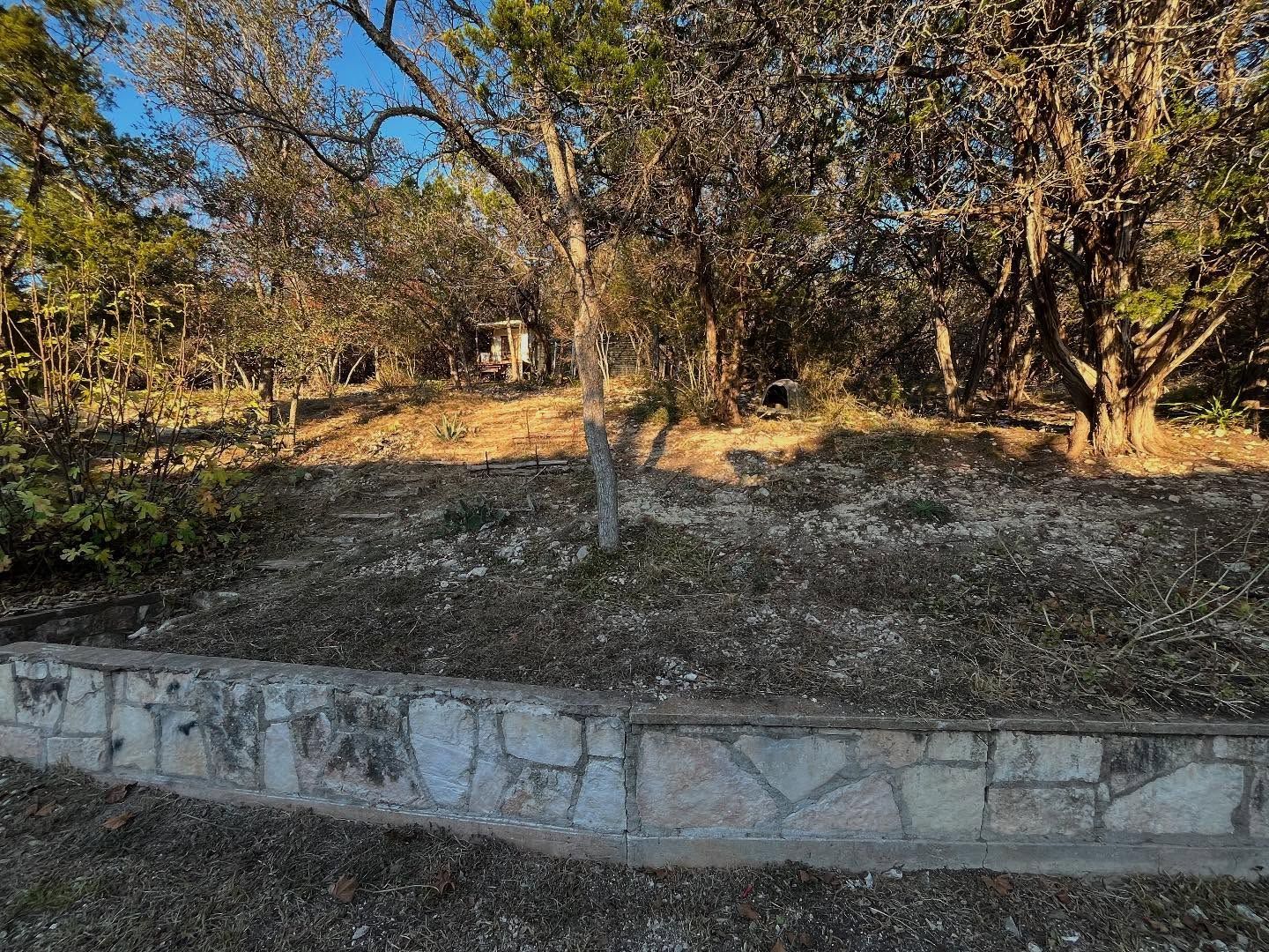 Stone retaining wall in front of a yard with trees, plants, and sunlight.
