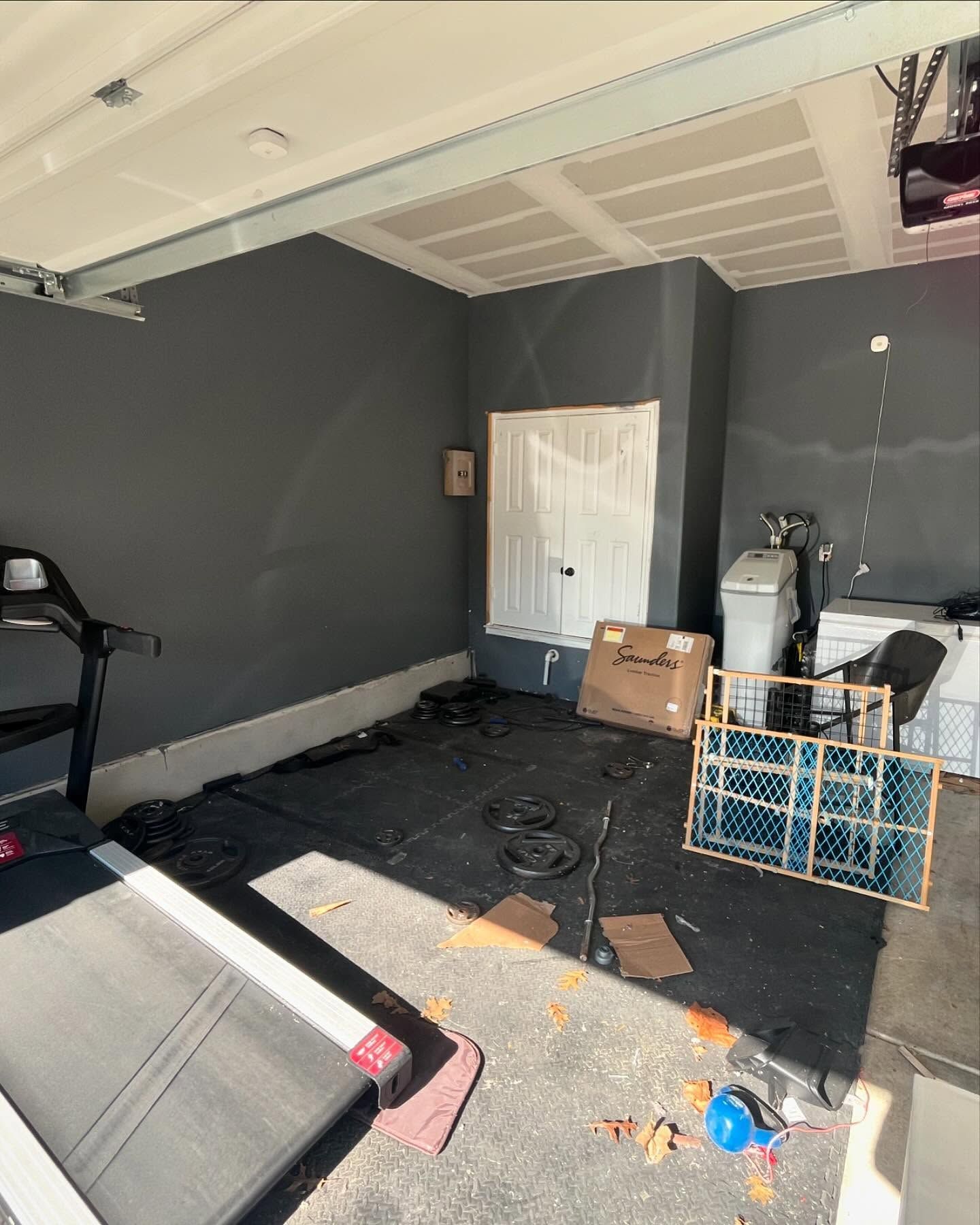 A partially finished garage with dark gray walls. A treadmill is in the foreground. Various items and debris litter the floor.