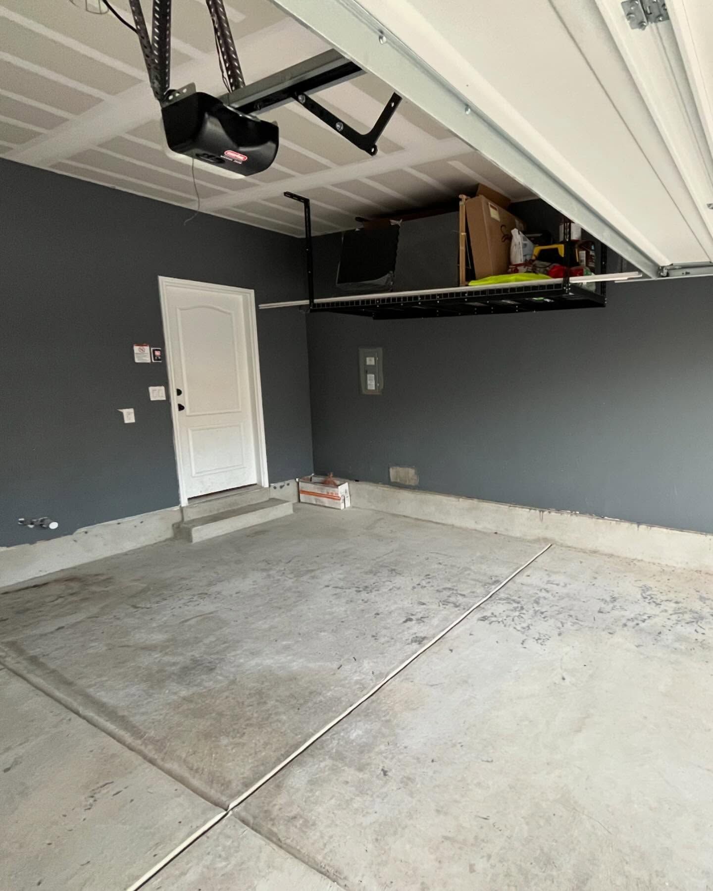 Gray garage interior with a concrete floor, a white door, and storage shelving near the ceiling.