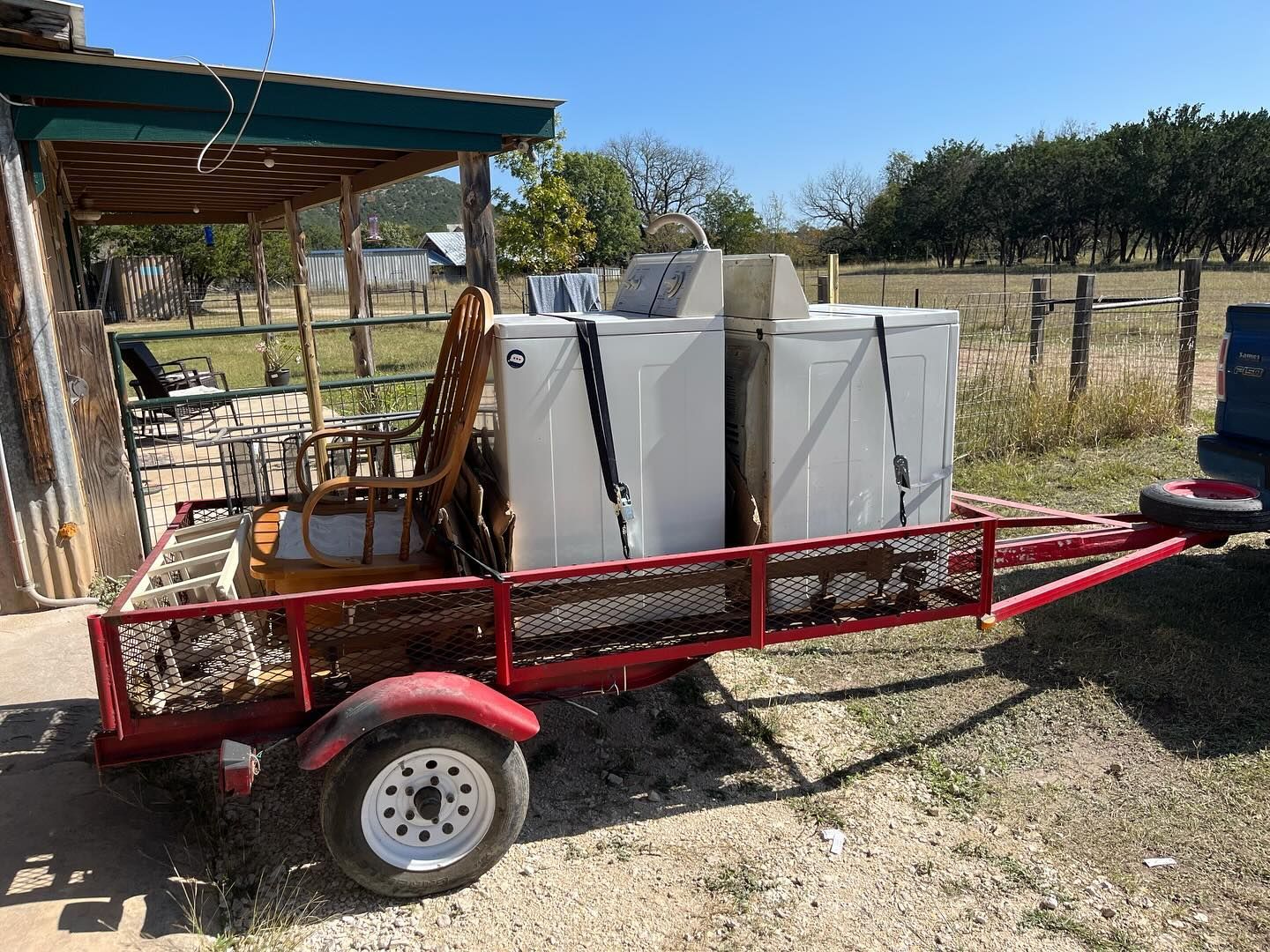 Red trailer loaded with white appliances, tied down. Chair also on trailer. Sunny, outdoor setting.