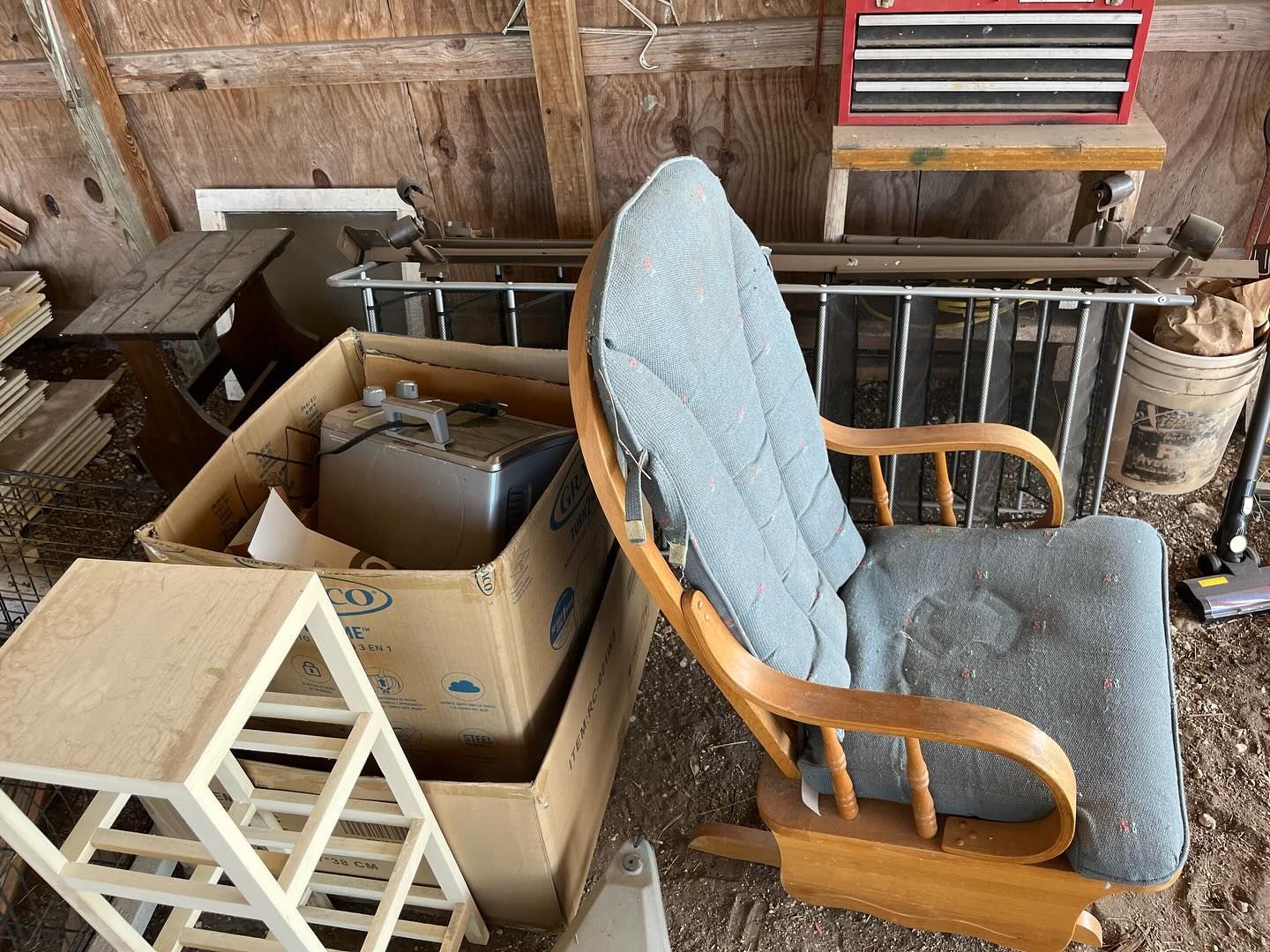 A cluttered wooden shed interior with a rocking chair, boxes, and a small wooden table.