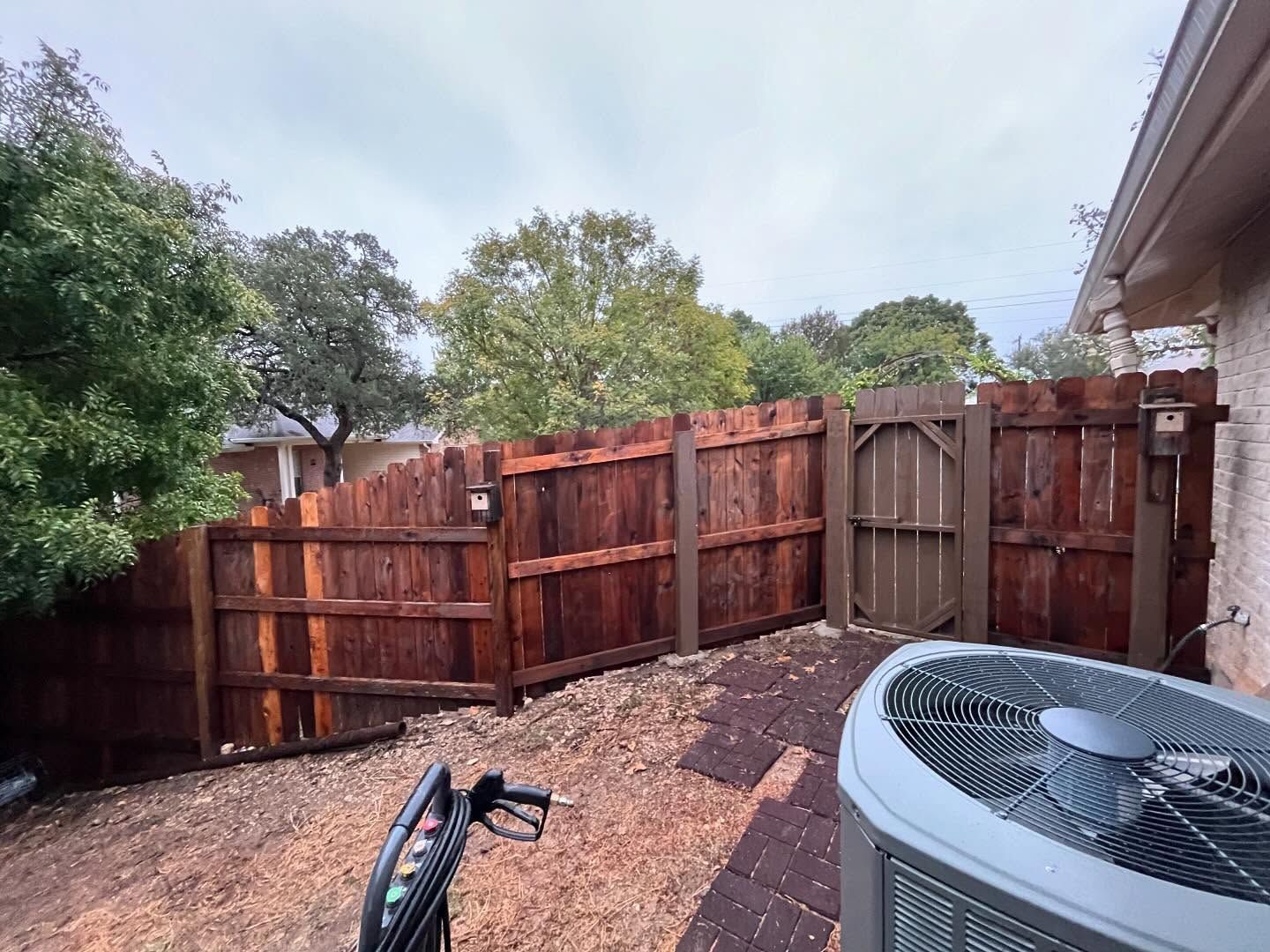 A backyard with a wooden fence, gate, and air conditioning unit. Overcast sky.