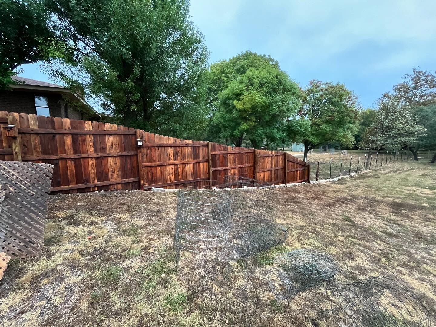 Wooden fence stained brown in a backyard with dry grass and trees under an overcast sky.